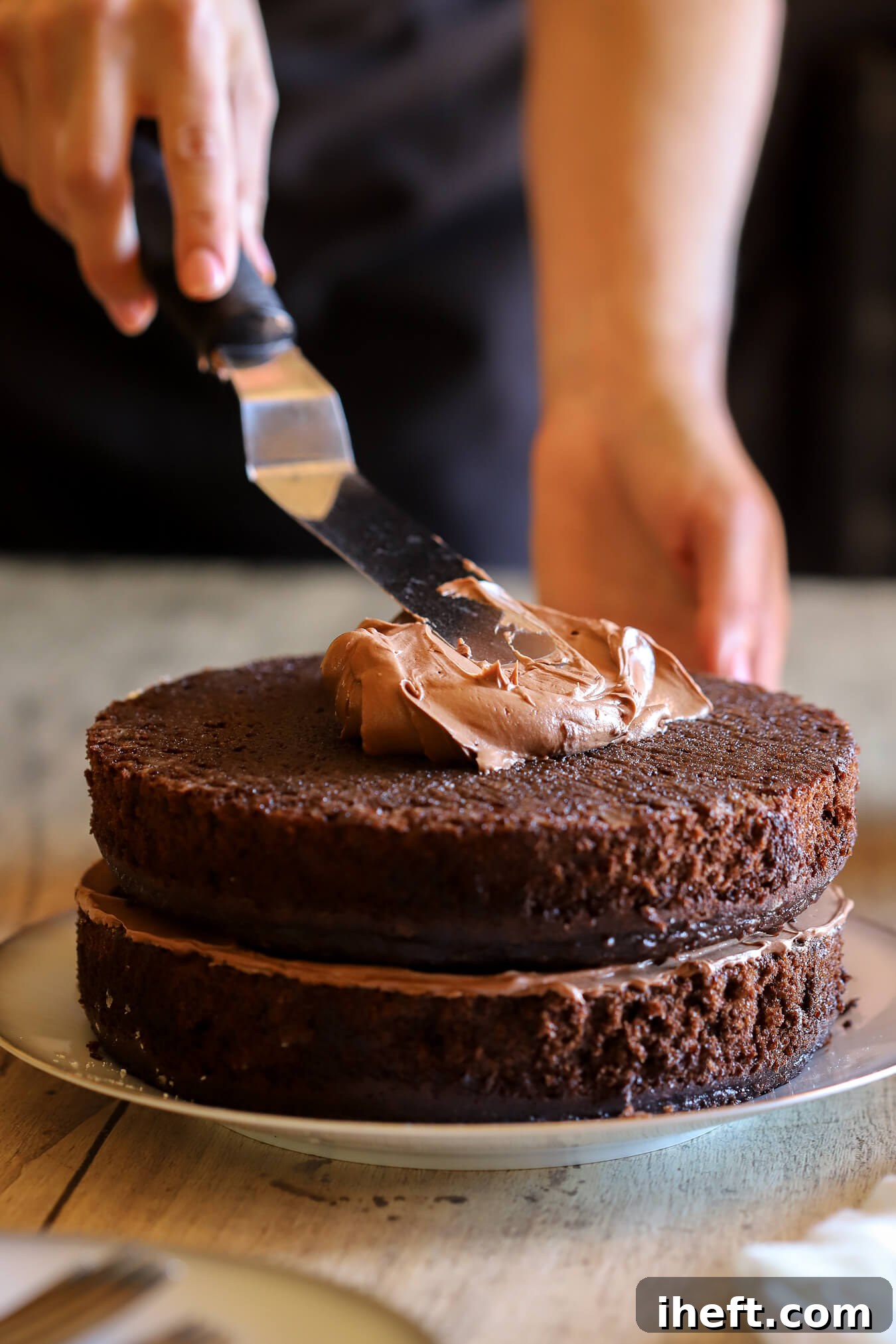 A skilled baker gracefully spreads rich chocolate buttercream onto a two-layer chocolate cake using an offset spatula, creating a smooth and even coating. This essential step transforms the baked layers into a beautifully frosted dessert.