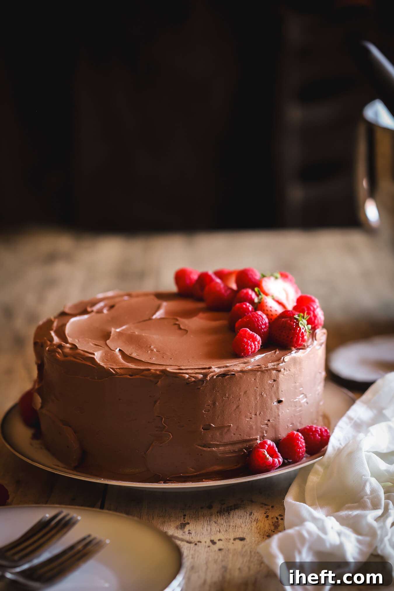 A stunning double chocolate Beatty's Chocolate Cake, derived from Ina Garten's Barefoot Contessa at Home cookbook, is elegantly presented on a wooden table against a dark, dramatic background. This two-layer chocolate cake is beautifully garnished with an assortment of fresh berries, enhancing its decadent appeal.