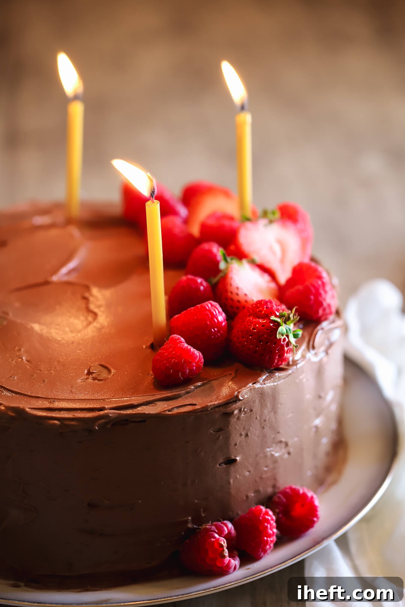 A captivating close-up of a two-layer chocolate birthday cake, adorned with glowing lit candles and a scattering of fresh berries for garnish. This inviting image highlights the cake's festive and delicious appeal.