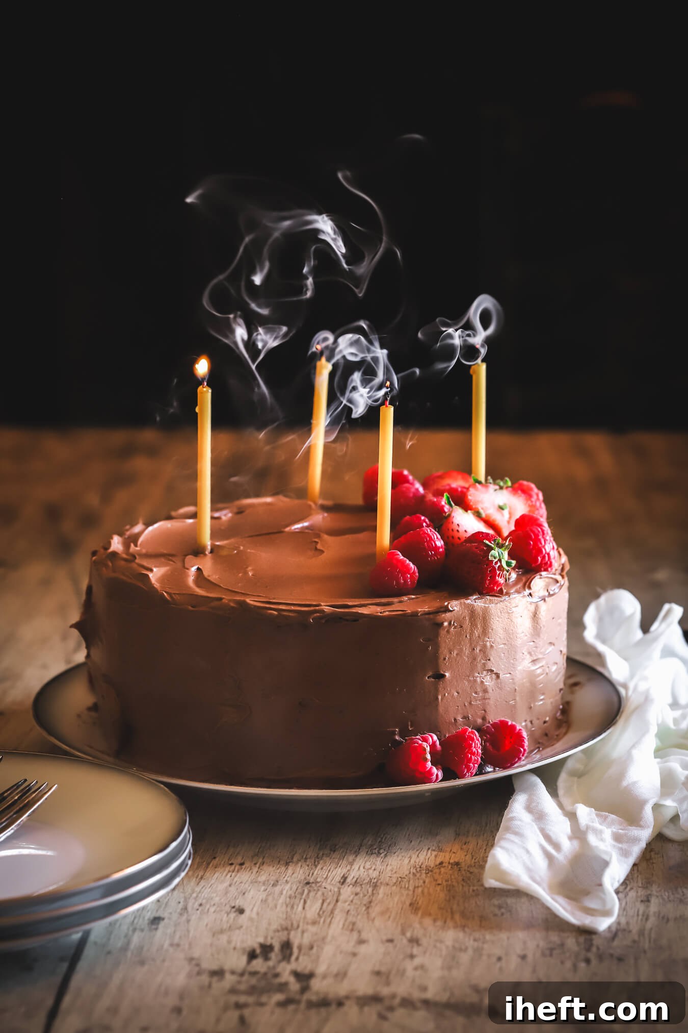 Beautiful food photography showcasing a double chocolate layer cake, featuring rich cocoa chocolate cake and silky chocolate buttercream frosting. The cake is adorned with four recently blown-out candles and garnished with fresh berries. This is Ina Garten's celebrated recipe for 