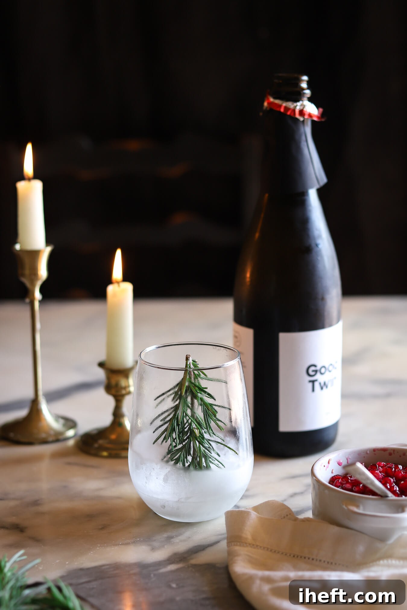 A frosty glass with a frozen rosemary sprig christmas tree sits on a marble table with a bottle of Champagne and candles in the background and bowl of pomegranate seeds in the foreground. The making of a snow globe Christmas Cocktail. 