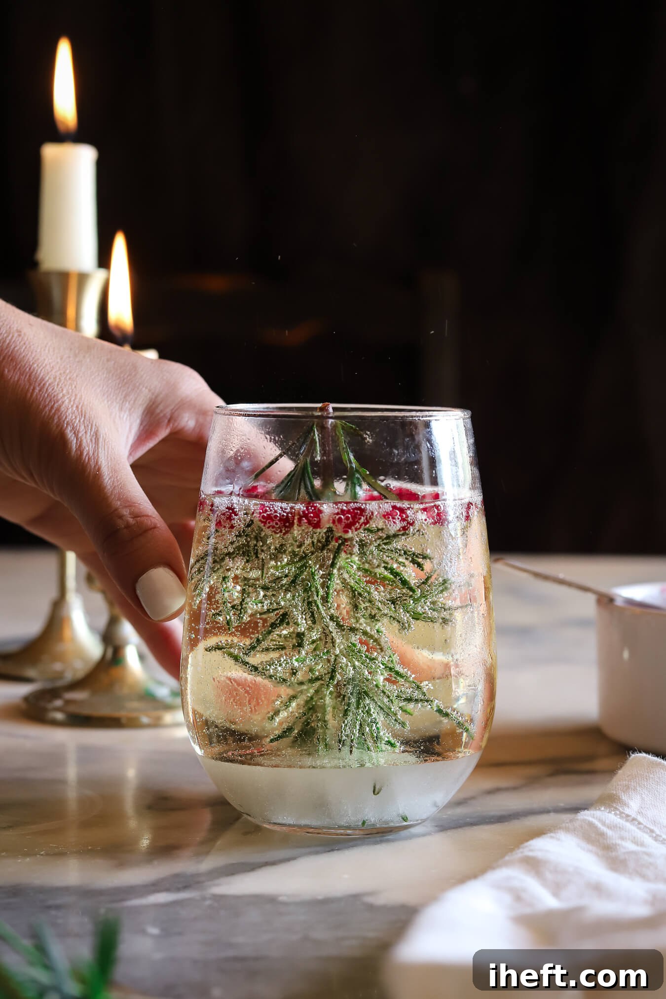 A bubbly snow globe Christmas cocktail with a rosemary christmas tree and pomegranate seeds on a marble table with hand in frame photography. 