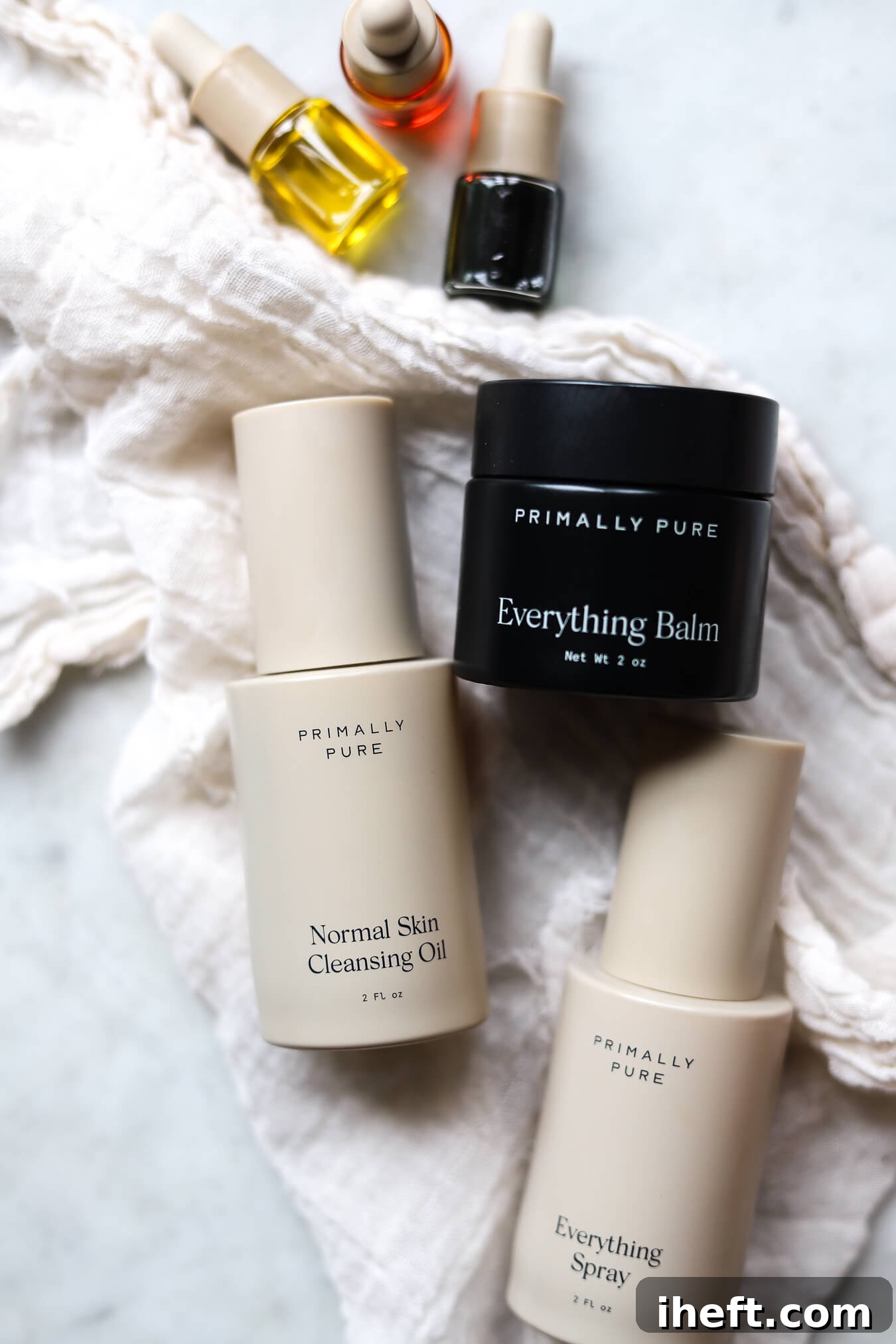 A collection of Primally Pure non-toxic skincare products, including three serums, Everything Balm, Cleansing Oil, and Everything Spray, arranged aesthetically on a white marble countertop.