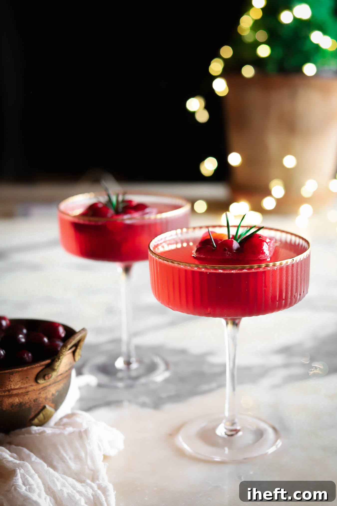 Beautiful food photography of two coupe glasses with gold rims filled with a red holiday mocktail drink with large cranberry ice cubes in the middle of the drinks. They sit on a marble counter with sparkly small potted tree in the background. 