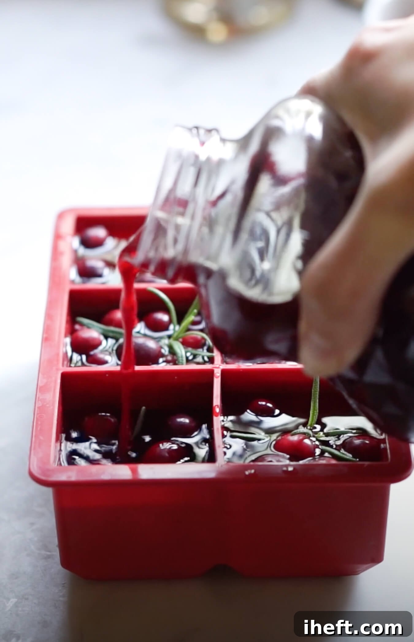 Cranberry juice is poured into a red silicon large ice cube mold with cranberries and rosemary to make cranberry ice.