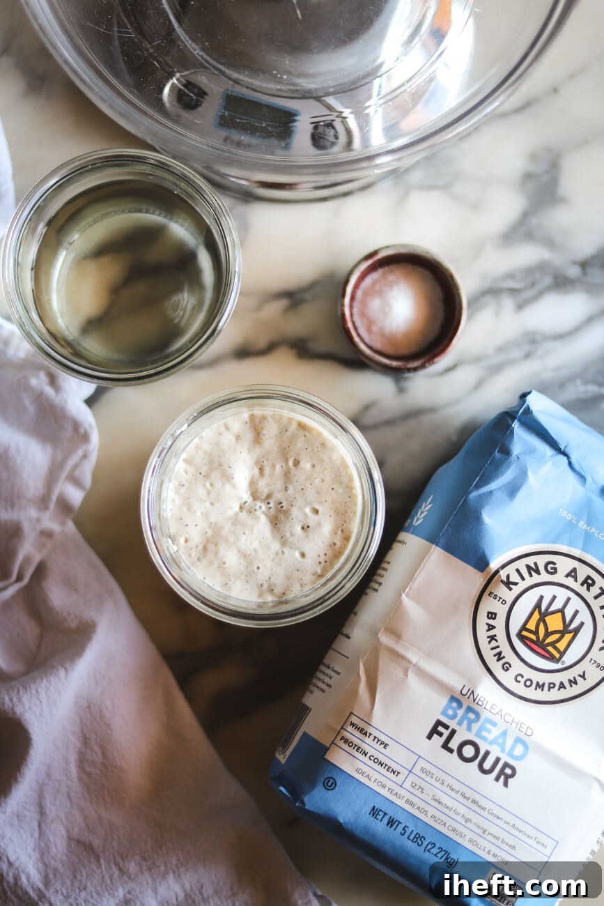 An overhead photo of the ingredients for homemade sourdough bread: a kitchen scale with bowl, salt, King Arthur bread flour, active sourdough starter, and water.