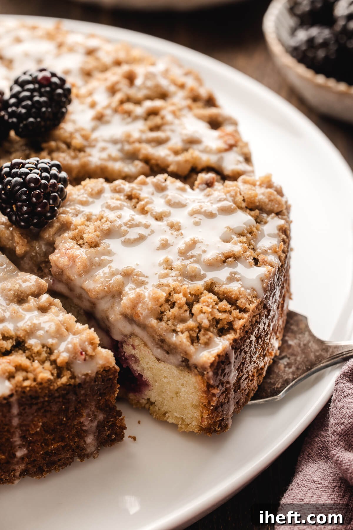 A serving fork lifts a portion of sour cream coffee cake, revealing its moist interior and crumbly streusel topping, against a backdrop of a white serving plate.