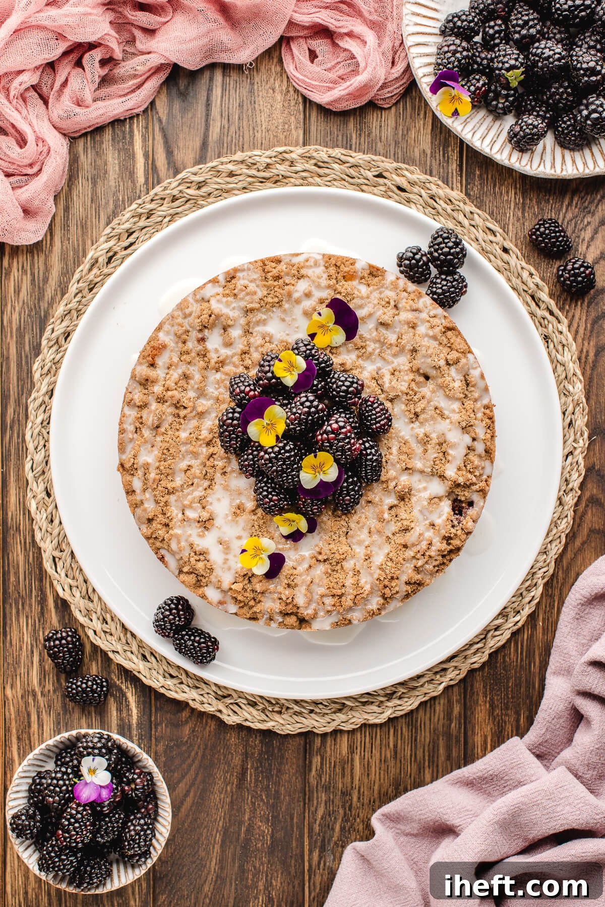A beautiful, whole blackberry coffee cake, elegantly garnished with fresh blackberries and delicate edible viola flowers, rests on a white serving plate. This stunning breakfast cake is ideal for festive brunches like Easter or Mother's Day, showcasing its seasonal appeal.