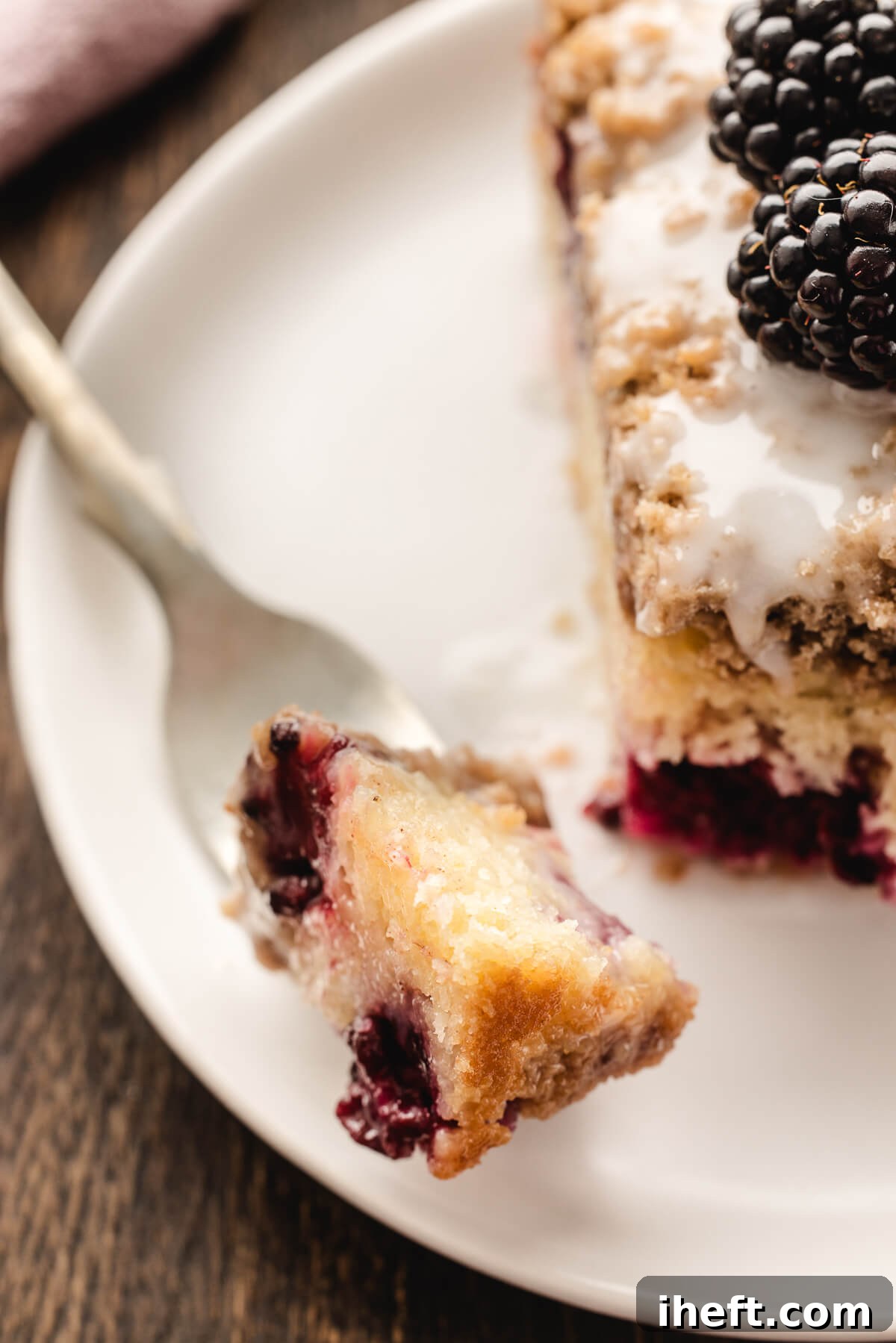 A close-up food photograph of a piece of blackberry sour cream coffee cake on a fork, poised above a white plate with a full slice of the cake, highlighting its moist crumb, streusel, and berry filling.
