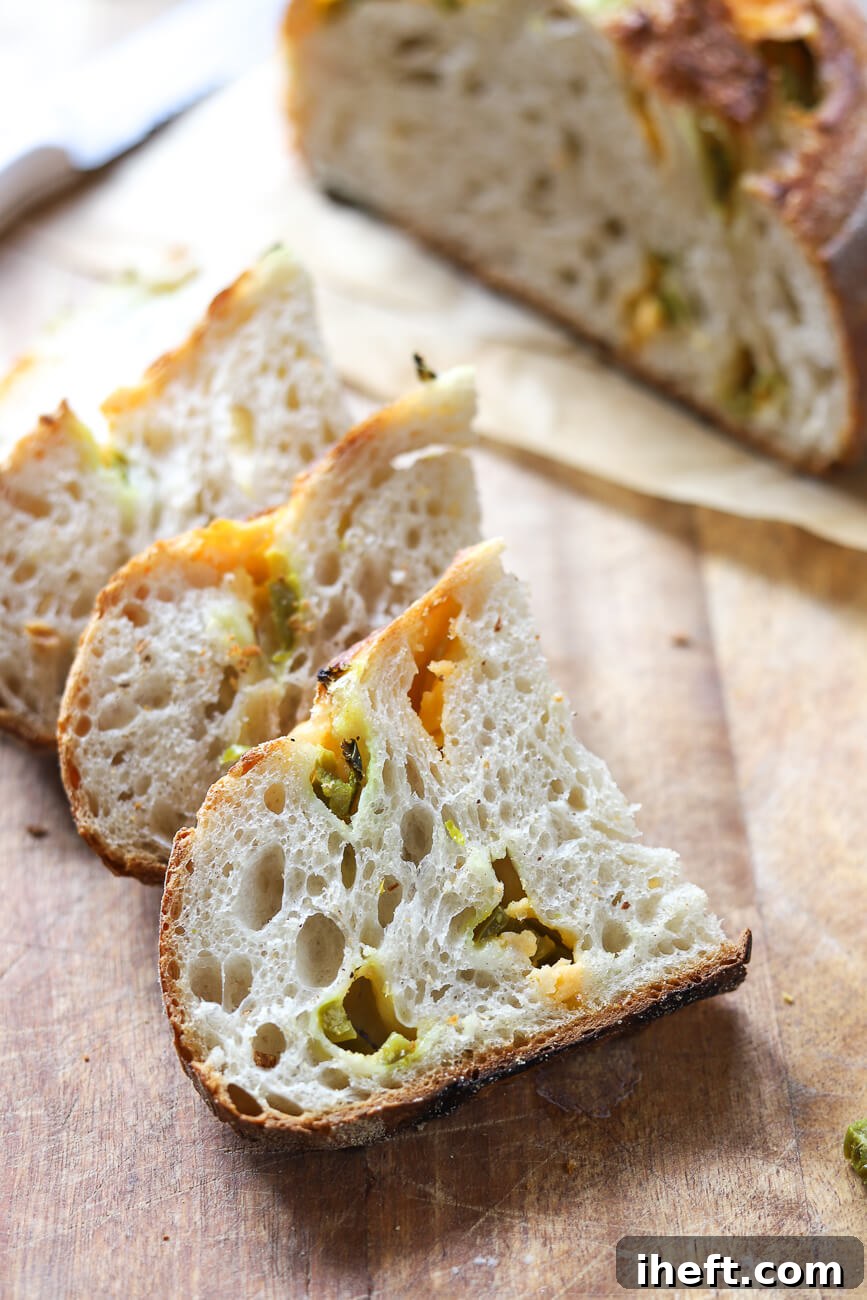 Slices of jalapeno cheddar sourdough on a cutting board with half the loaf in the background. This bread has an open crumb and is dotted with pickled sliced jalapenos and cheddar cheese. 