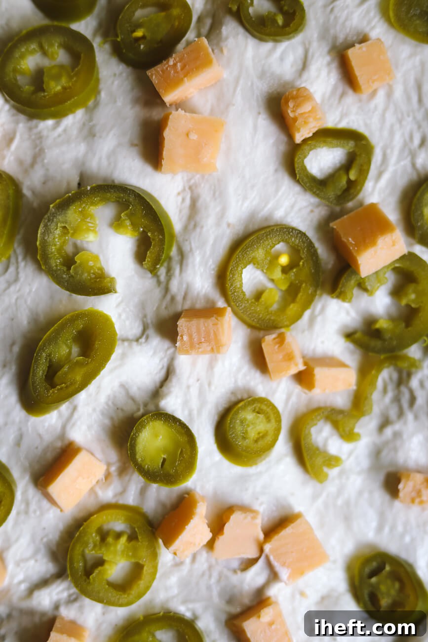 A close-up photo of cheddar cheese cubes and sliced jalapeno peppers on sourdough bread dough before folding in. This shows how to add inclusions to sourdough. 