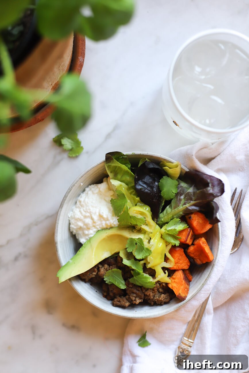 A high protein ground beef, roasted sweet potato bowl that is viral on Tiktok. This one sits on a marble counter next to a glass of ice water and is topped with cilantro, pepperoncini, and greens with a scoop of cottage cheese. 