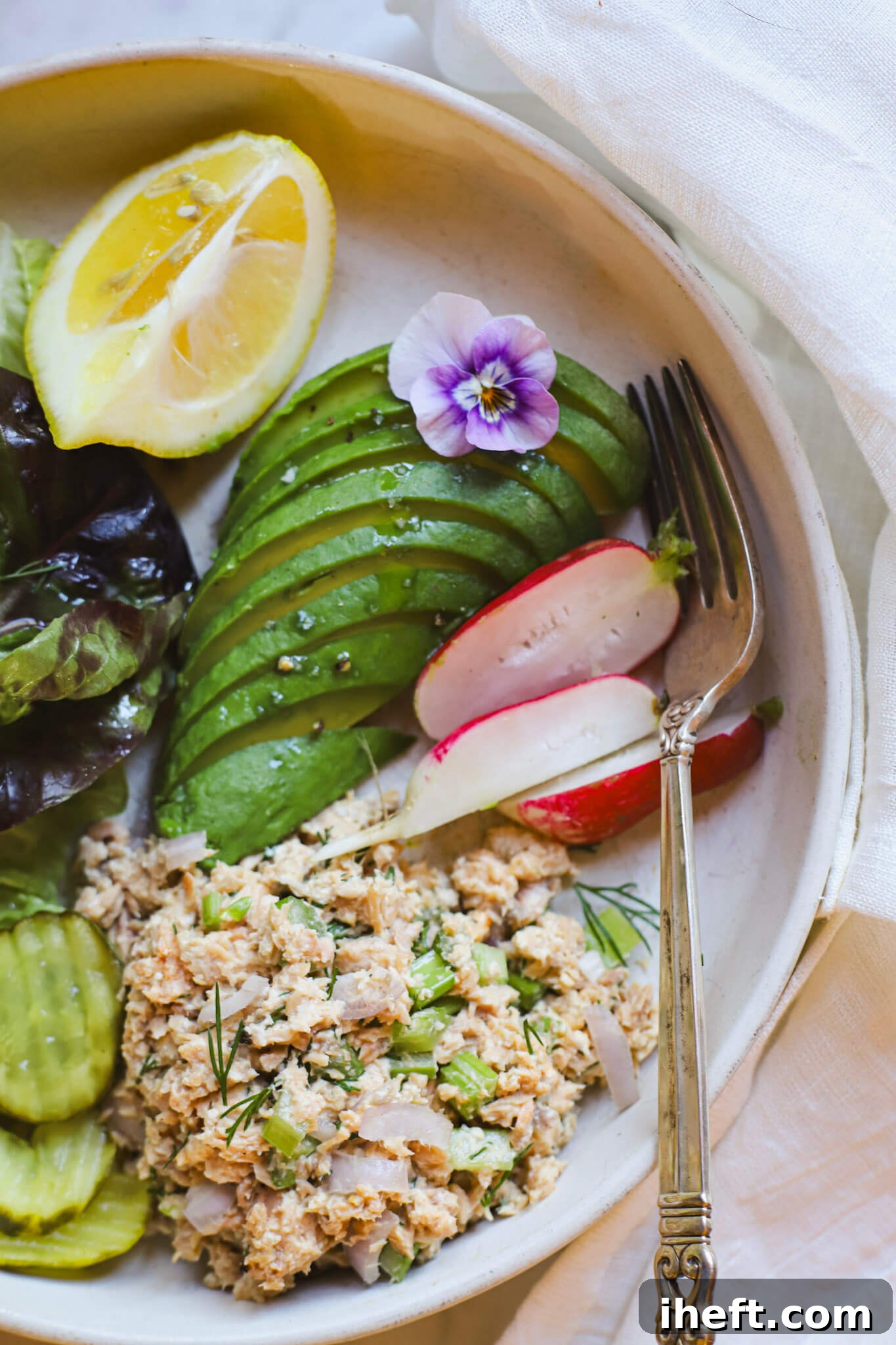 A scoop of salmon salad made with canned salmon, celery, onion, mayo and dill on a plate with sliced avocado, radishes, pickles, lemon and greens for a high protein lunch.