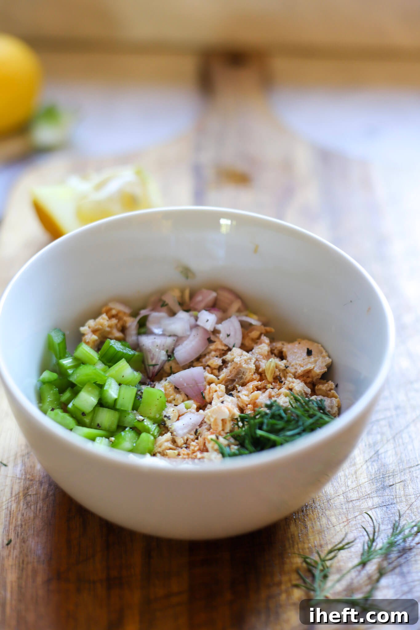 A small white ceramic bowl filled with canned salmon, dill, onion, and celery before being mixed together for a healthy high protein lunch.