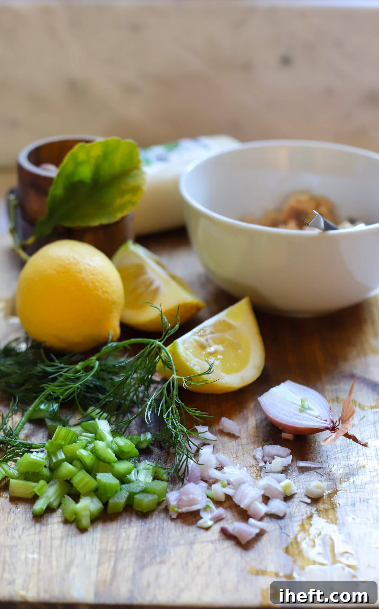 Diced celery, onions, dill, lemons, a salt cellar, mayo and canned salmon in a bowl sit on a wooden cutting board ready to make salmon salad.