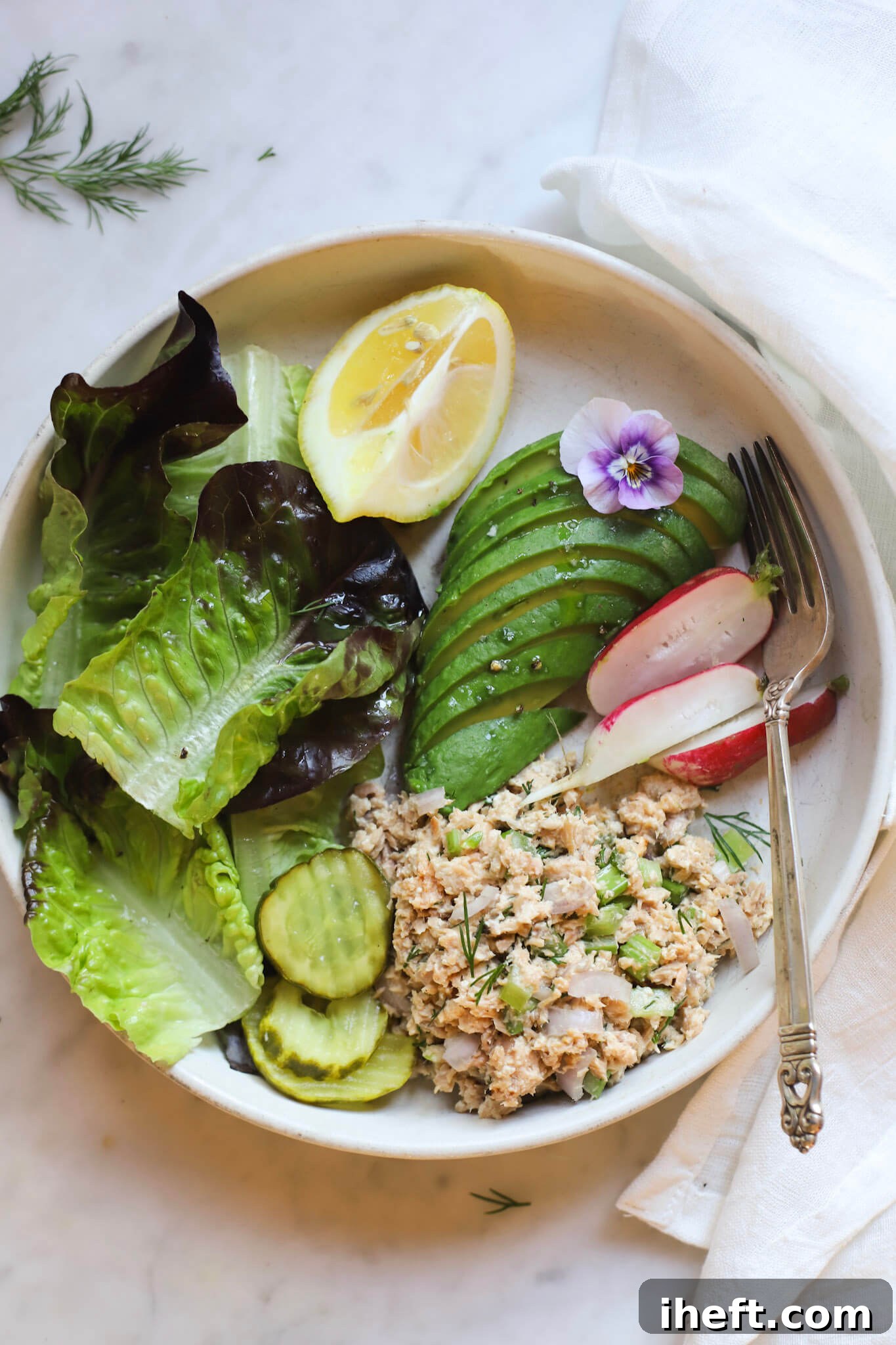 A delicious, easy, high protein, low carb lunch on a white ceramic plate: canned salmon salad with celery and dill, sliced pickles, greens, sliced avocado, and radish, garnished with a lemon wedge and edible flower.