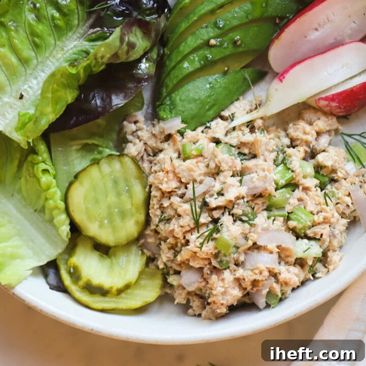 Canned salmon salad made with celery, dill, onion, and mayo on a plate with greens, avocado, radish, and pickles for a healthy lunch.