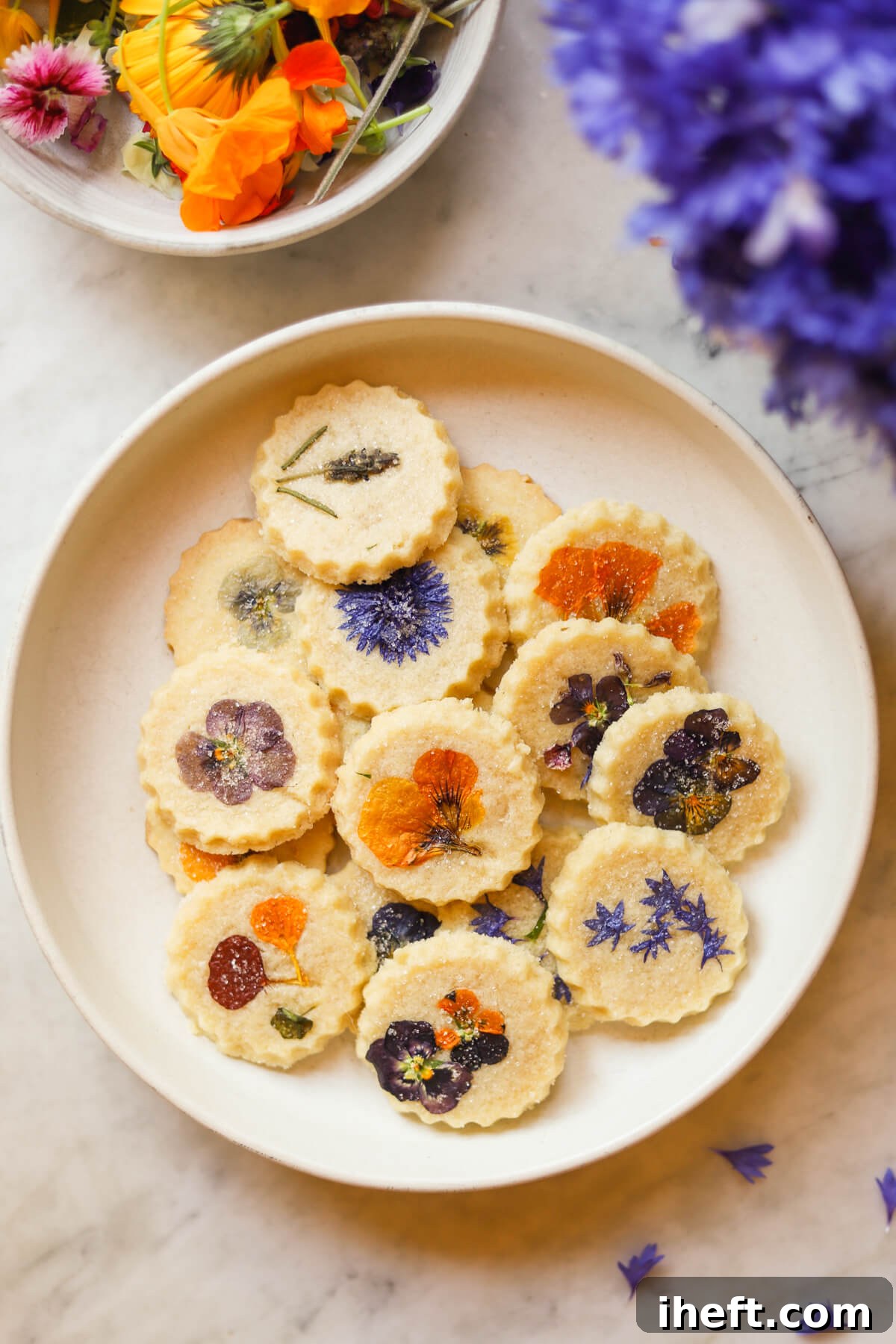 Beautiful food photography of a white ceramic plate of round shortbread cookies topped with pressed edible flowers. More fresh edible flowers are off to the sides.