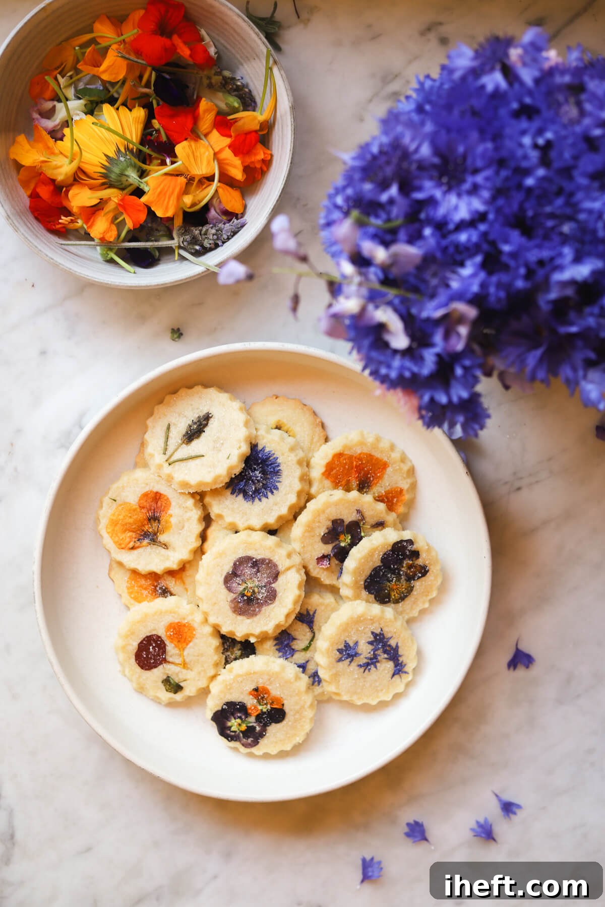 A bowl of edible flowers, a bouquet of purple Bachelor's buttons, and a plate of edible flower pressed cookies on a white plate on a marble counter.