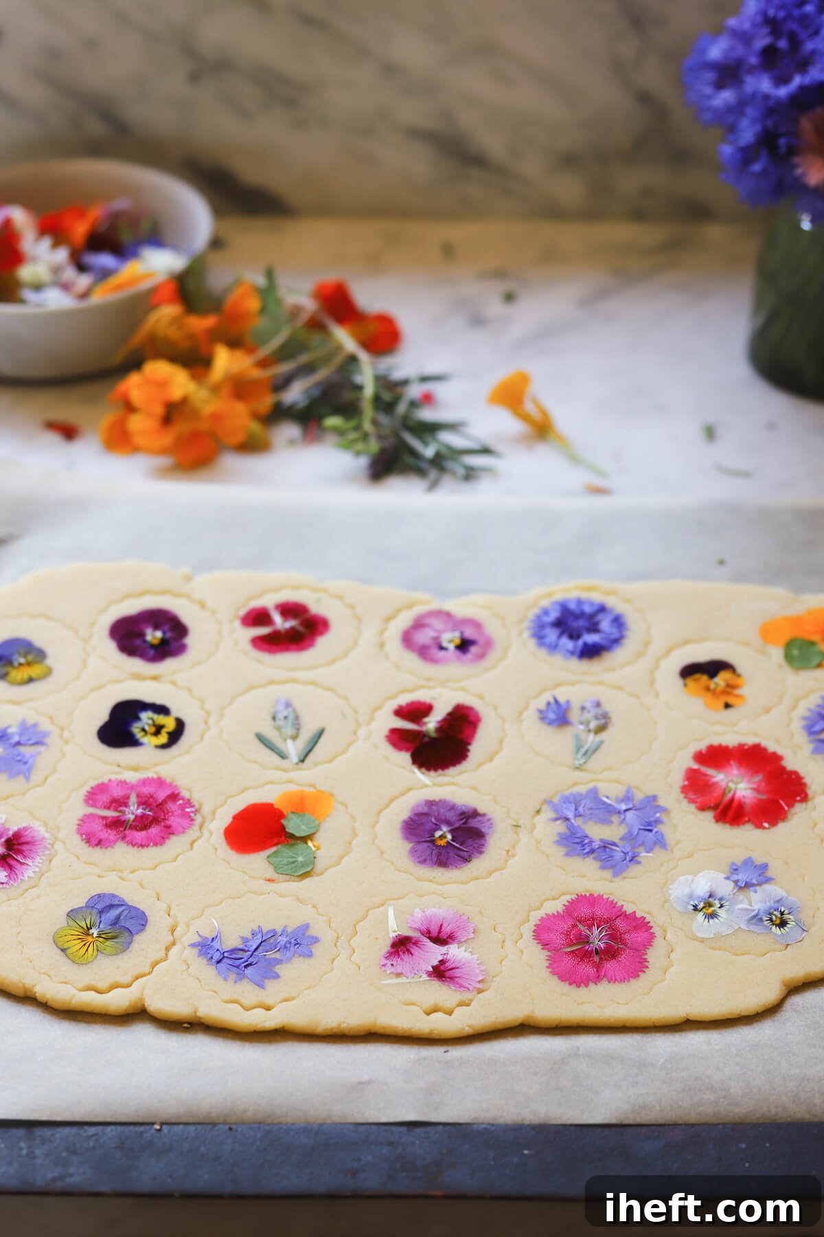 A sheet of shortbread cookie dough cut into circles and topped with edible flowers on a marble kitchen counter.