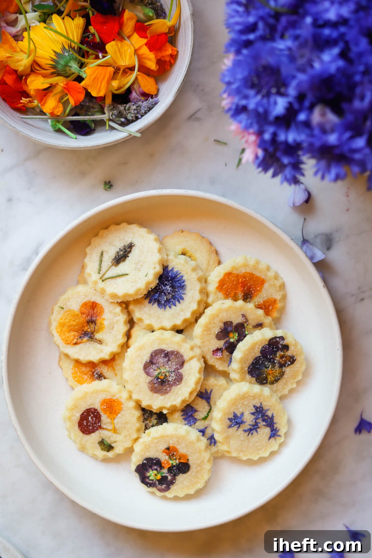 A plate of beautiful edible flower pressed shortbread cookies on a white plate next to Bachelor's buttons and other edible flowers including violas, lavender, and nasturtiums.