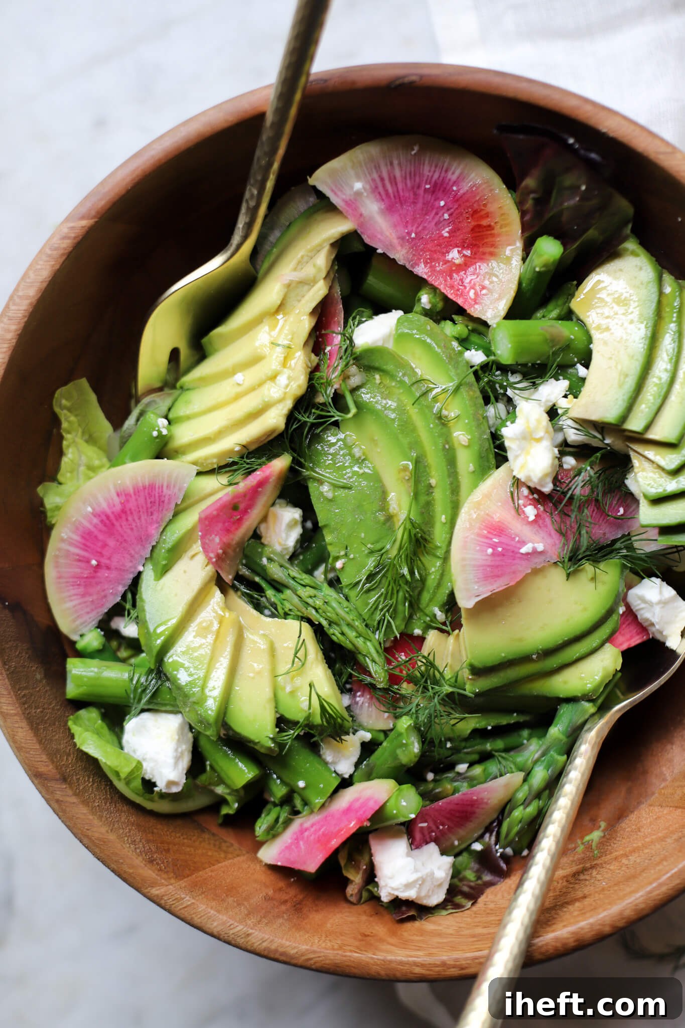 A beautiful spring salad made with blanched asparagus, sliced watermelon radish, crumbled feta, dill, and avocado in a wooden salad bowl with gold serving utensils. 