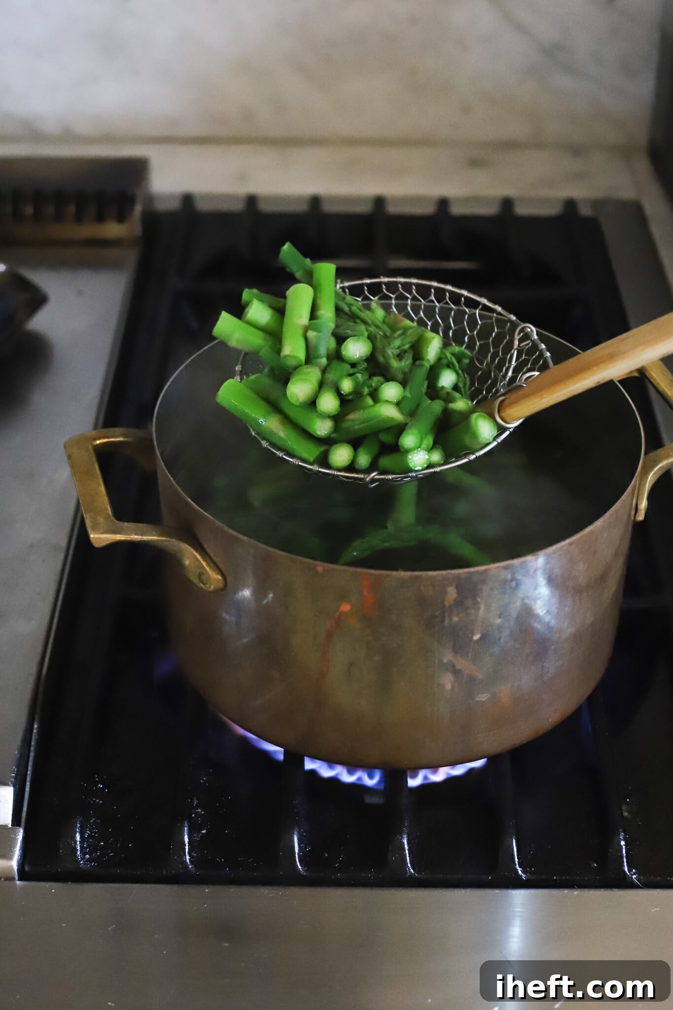 A slotted spoon lifts blanched asparagus pieces out of a copper pot of boiling water on a gas burner. 