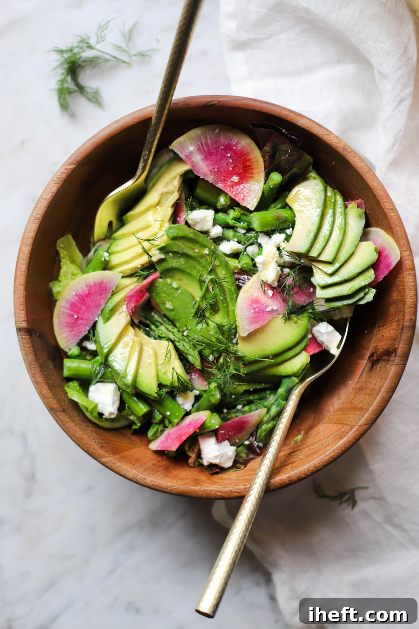 An overhead photo of a spring asparagus, avocado, feta, and radish salad in a wooden salad bowl with gold utensils on a white marble counter. 