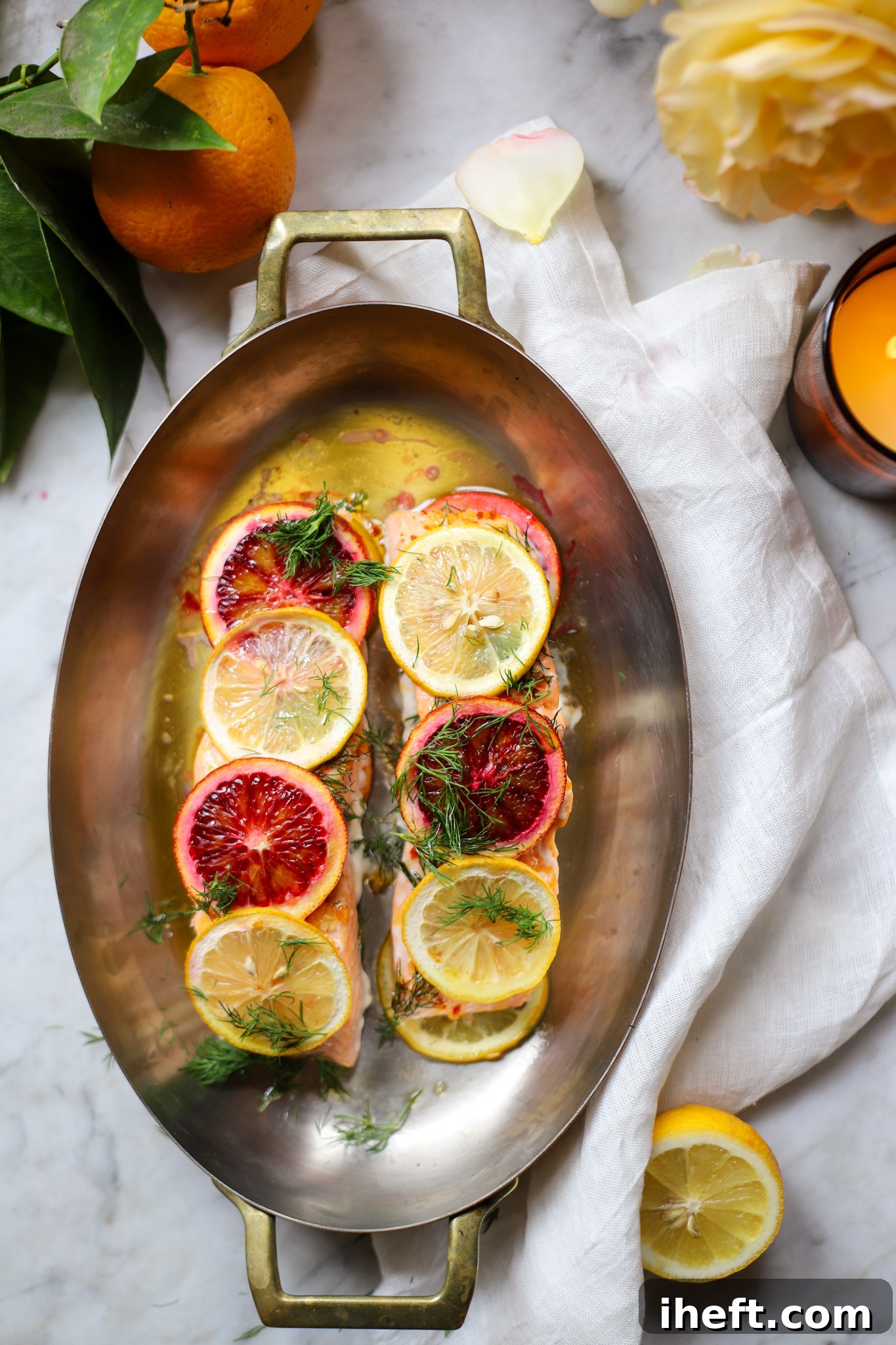 Overhead food photography of two salmon filets in an ovel baking dish. Salmon is topped with slices of blood orange and lemon and garnished with fresh dill. The dish sits on a white marble counter with white napkin. 