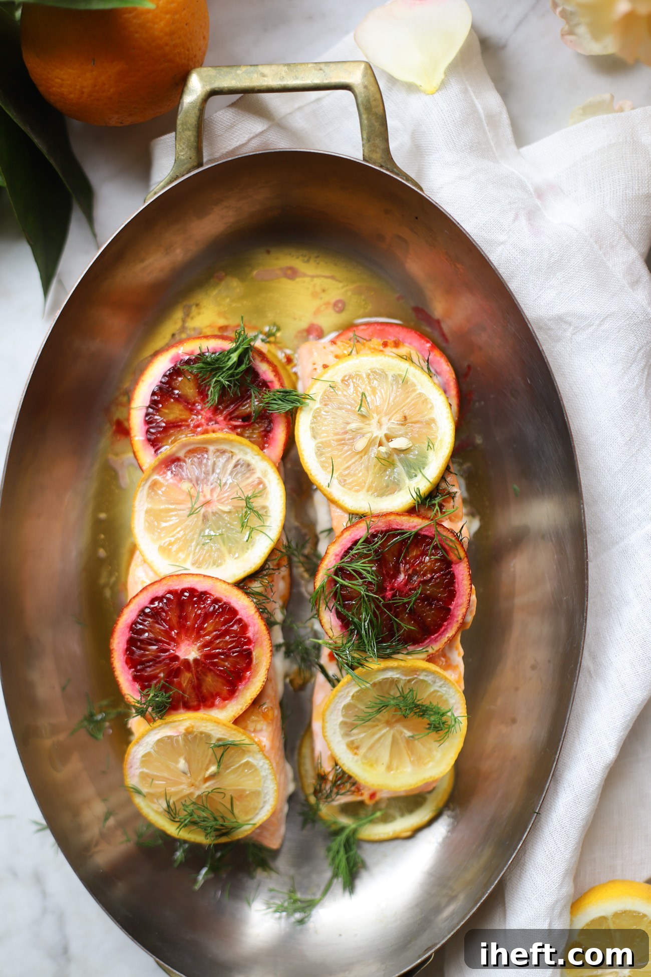 Salmon filets in an oval copper baking dish topped with orange and lemon slices and dill. 