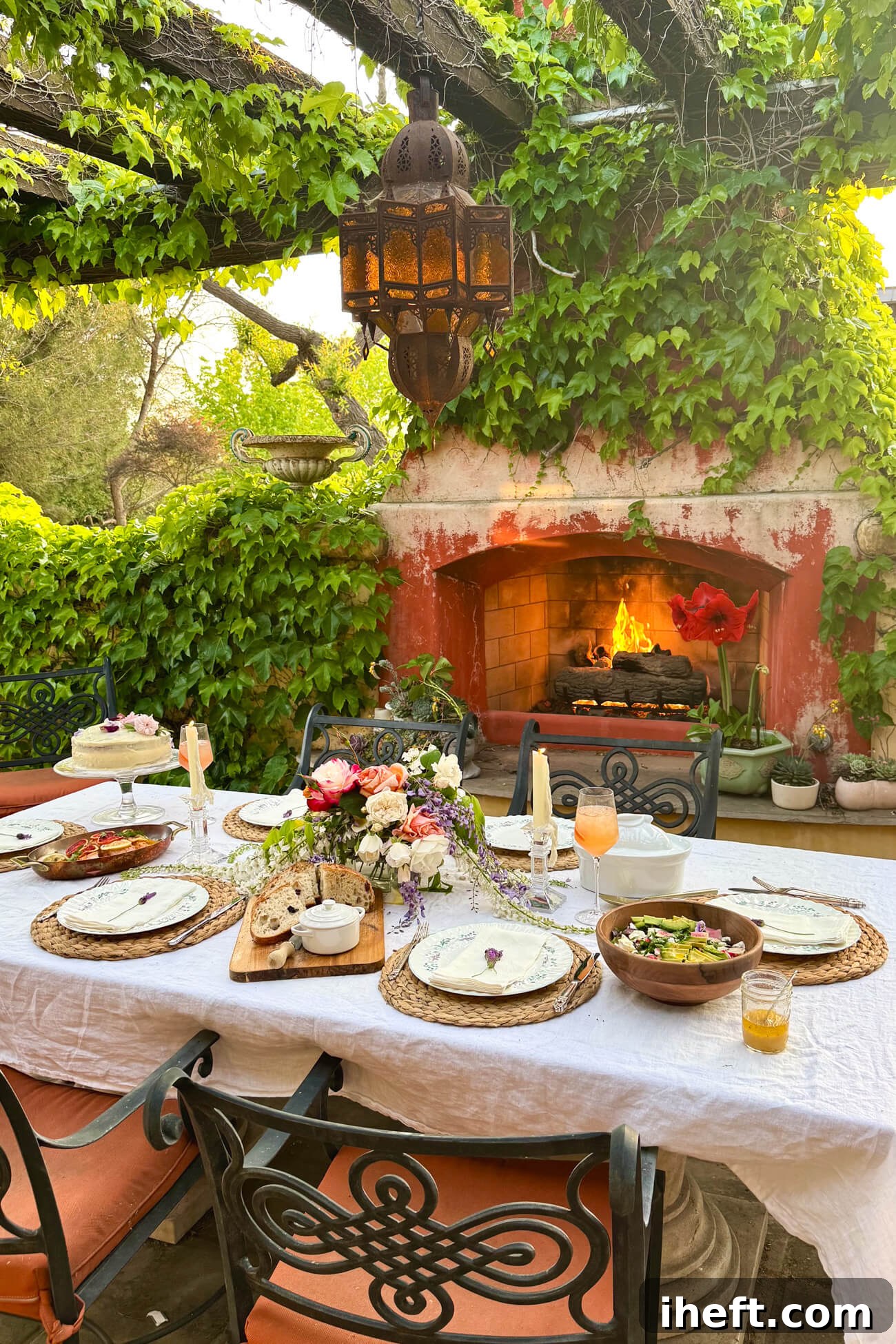 An outdoor table set with a spring flower arrangement in the center, white tablecloth, salad, cake, and other dishes. A vine-covered outdoor fireplace in the background.