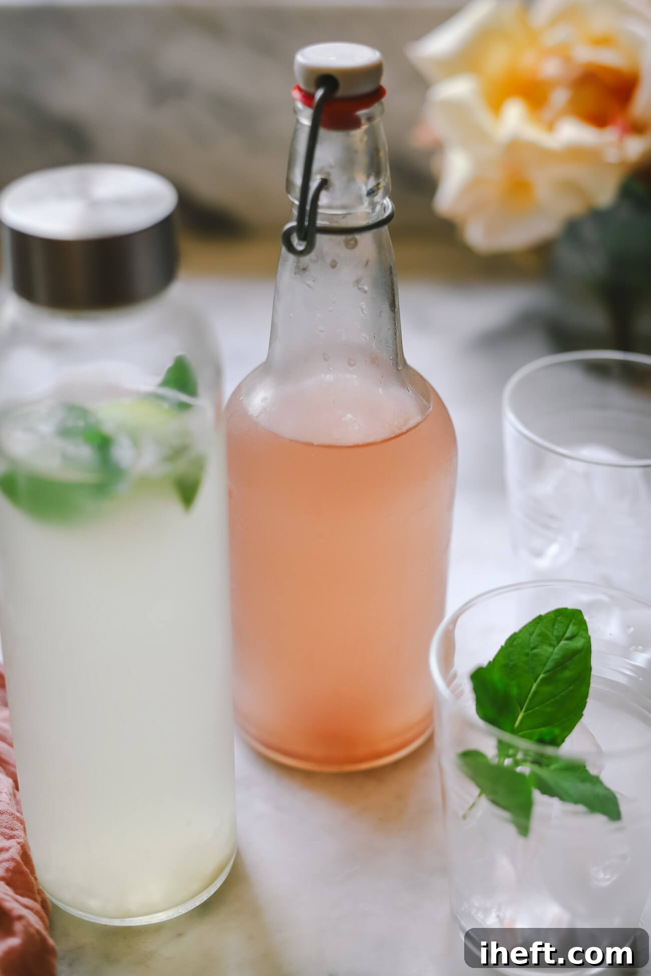 Two bottles of homemade water kefir during a second ferment. Flavors are mojito (coconut water, mint, lime) and pomegranate. The bottles sit on a marble countertop next to two glasses of ice. This homemade sparkling probiotic soda looks refreshing and delicious.