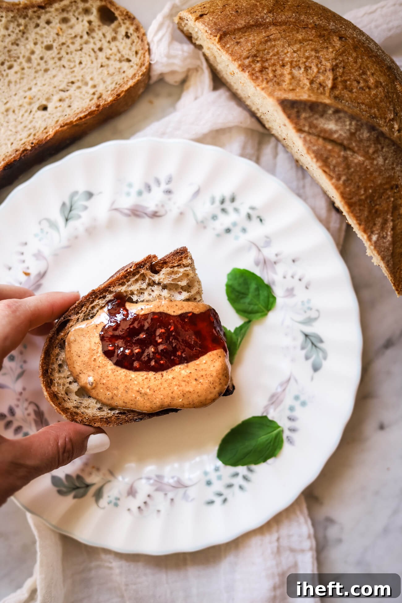 A hand lifts a half slice of gluten free sourdough bread topped with almond butter and raspberry jam. Half of the homemade boule sits off to the side. This delicious and healthy breakfast or snack is depicted through mouthwatering food photography. 