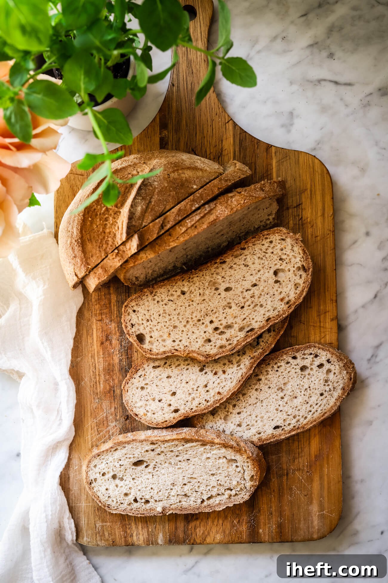 Beautiful overhead food photography of a loaf of homemade gluten free sourdough bread cut into slices on a wooden cutting board on a white marble counter. Fresh herbs and roses peek out from one corner. The bread slices look perfect for sandwiches or toast. 