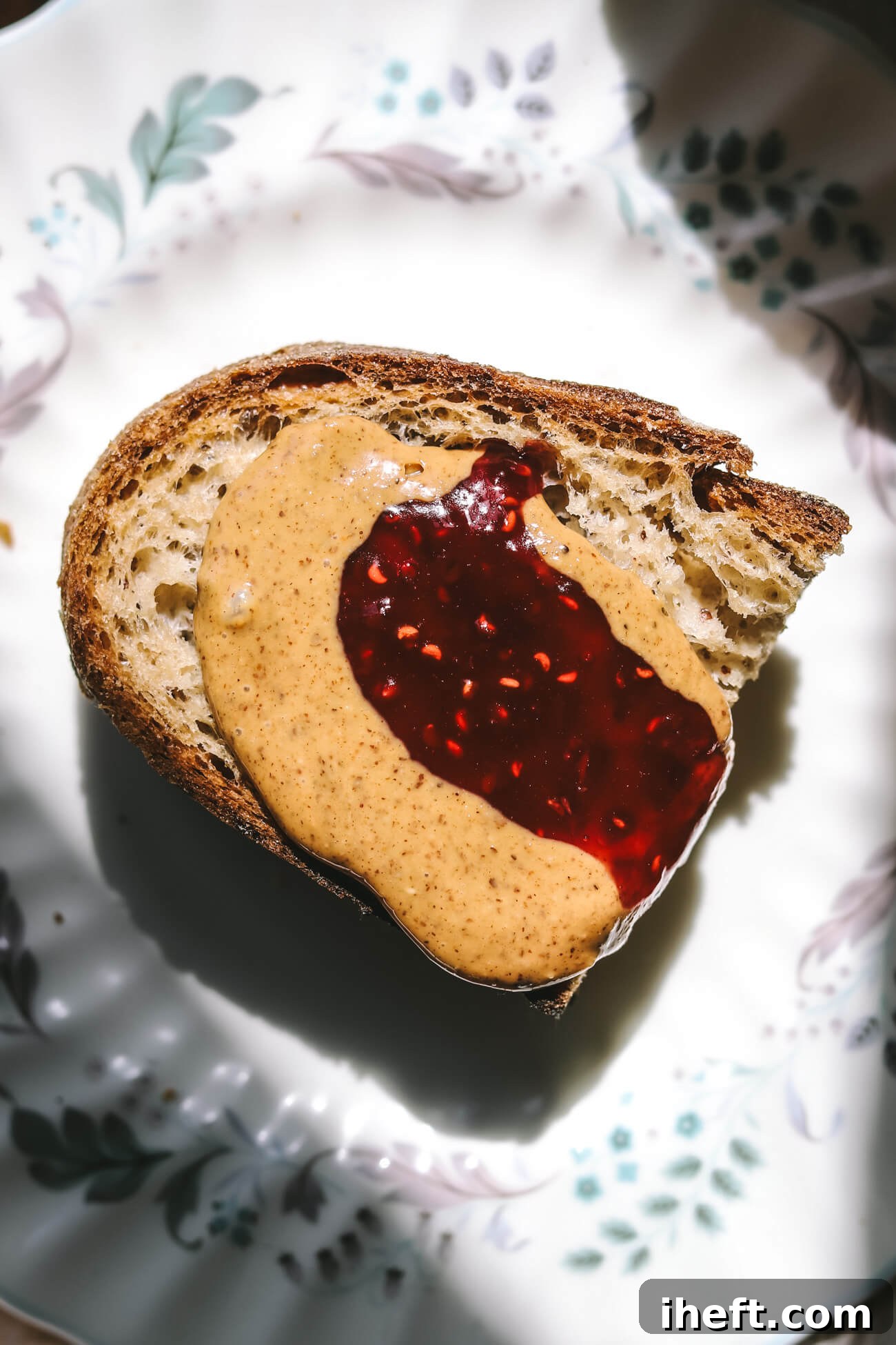 Close-up overhead photo half a slice of homemade gluten free sourdough toast topped with nut butter and raspberry jam on a delicate floral china plate. 
