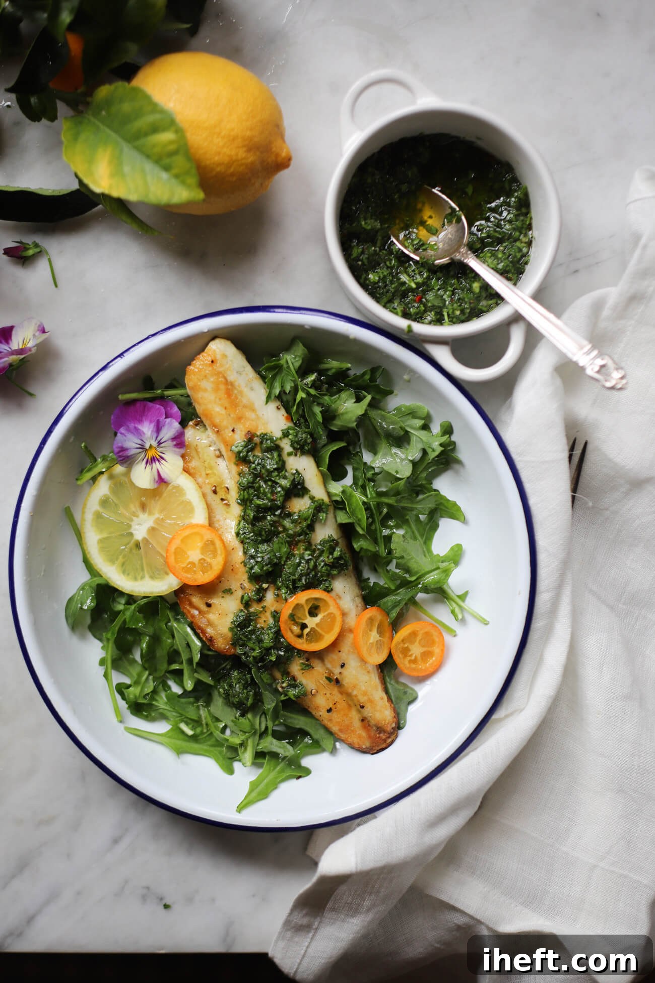 A beautifully plated branzino fillet, perfectly pan-seared with crispy skin, presented on a pristine white plate alongside fresh arugula, a side dish of vibrant gremolata, and bright kumquat slices. A lemon half adds a fresh touch, creating an appetizing and healthy meal.