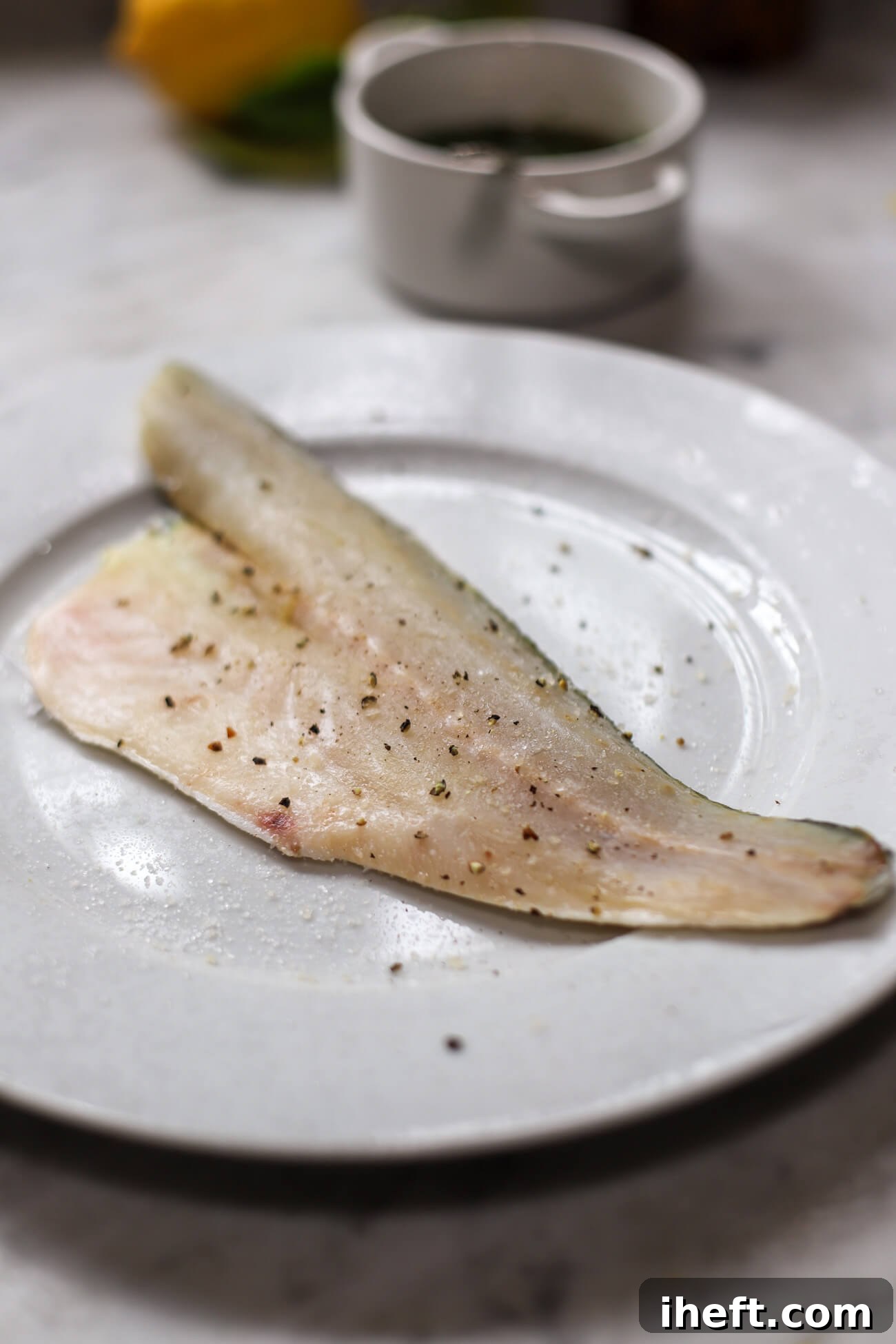 A large raw branzino fillet seasoned with salt and pepper on a white plate, ready for cooking.