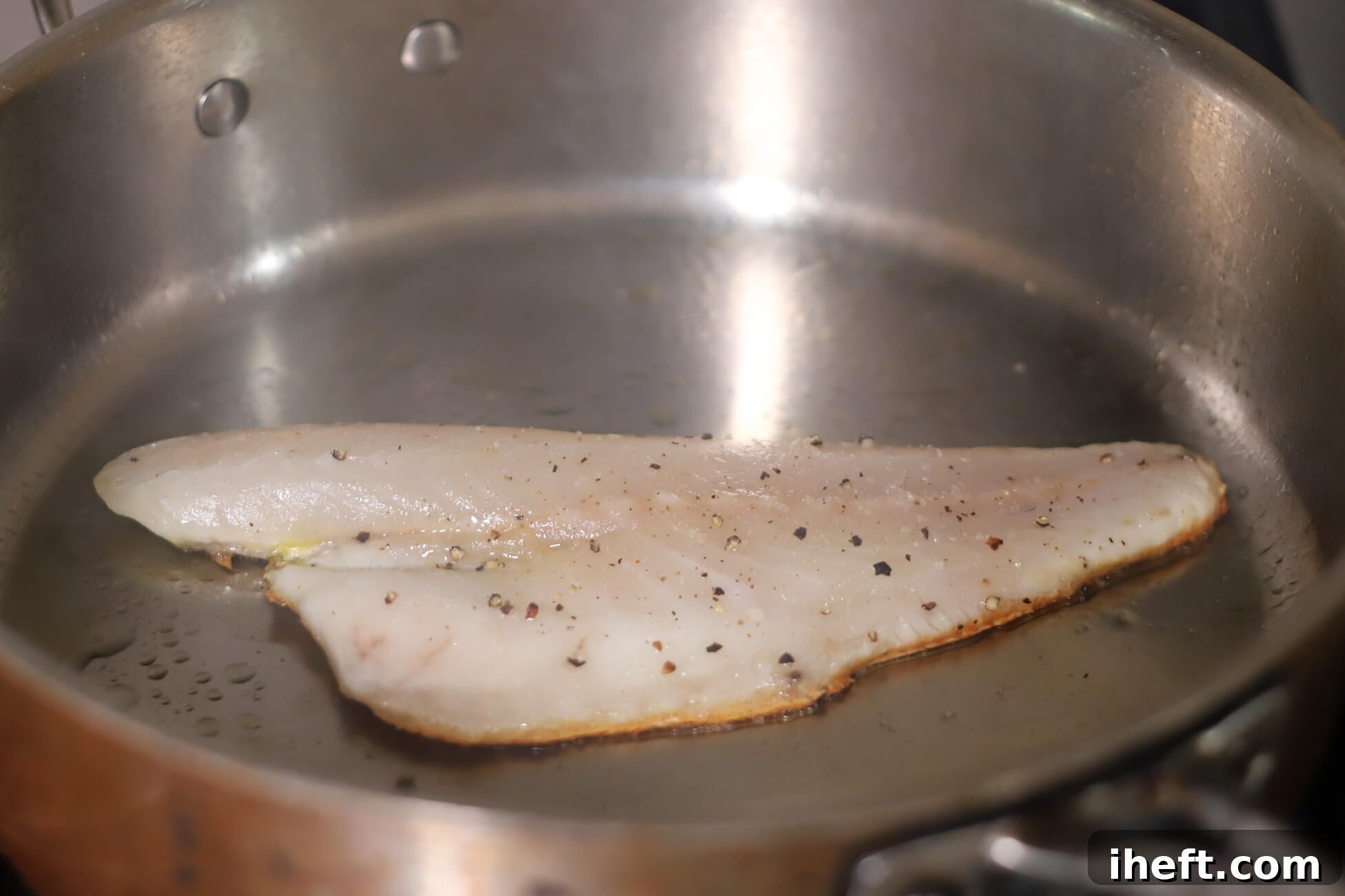 A branzino fillet pan-searing skin-side-down on a hot stainless steel skillet, achieving crispy skin.