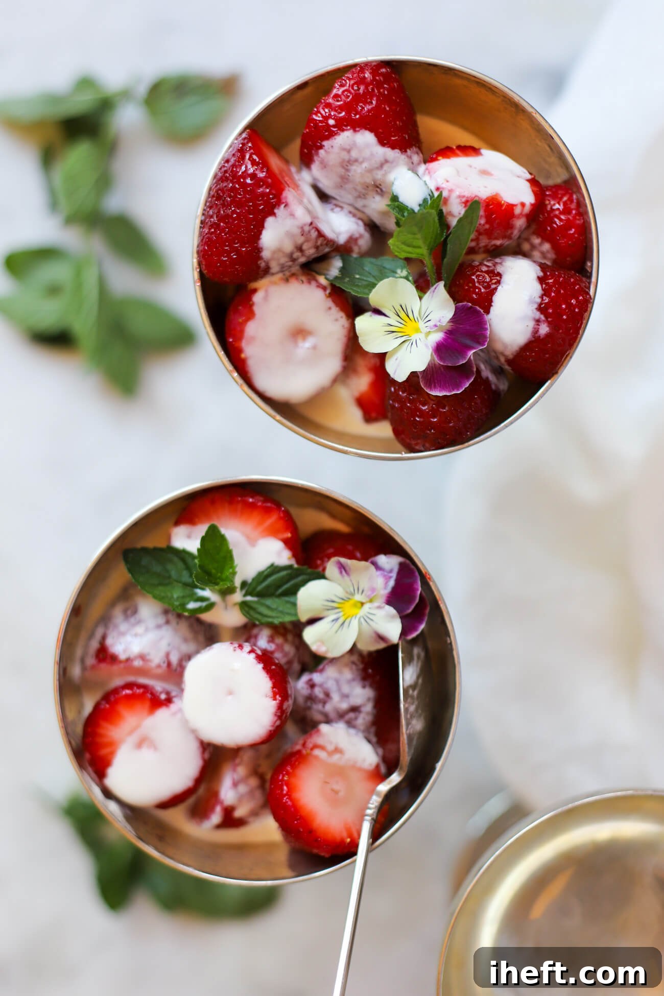 Overhead food photography of two bowls of Wimbledon-style strawberries and cream garnished with mint and violas. 