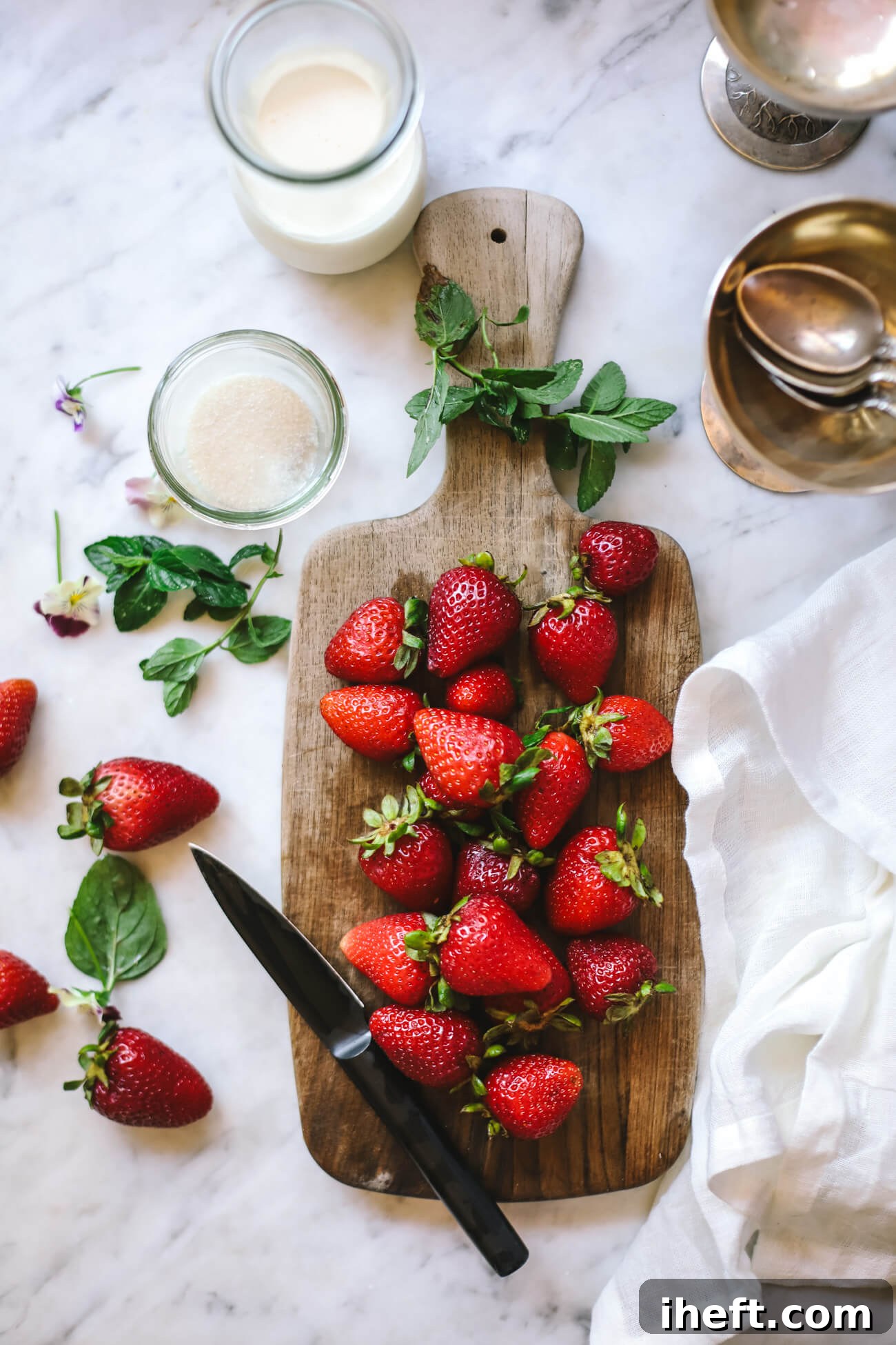 Overhead photo of strawberries on a wooden cutting board on a white marble counter alongside a small dish of granulated organic sugar and a small jar of cream. 