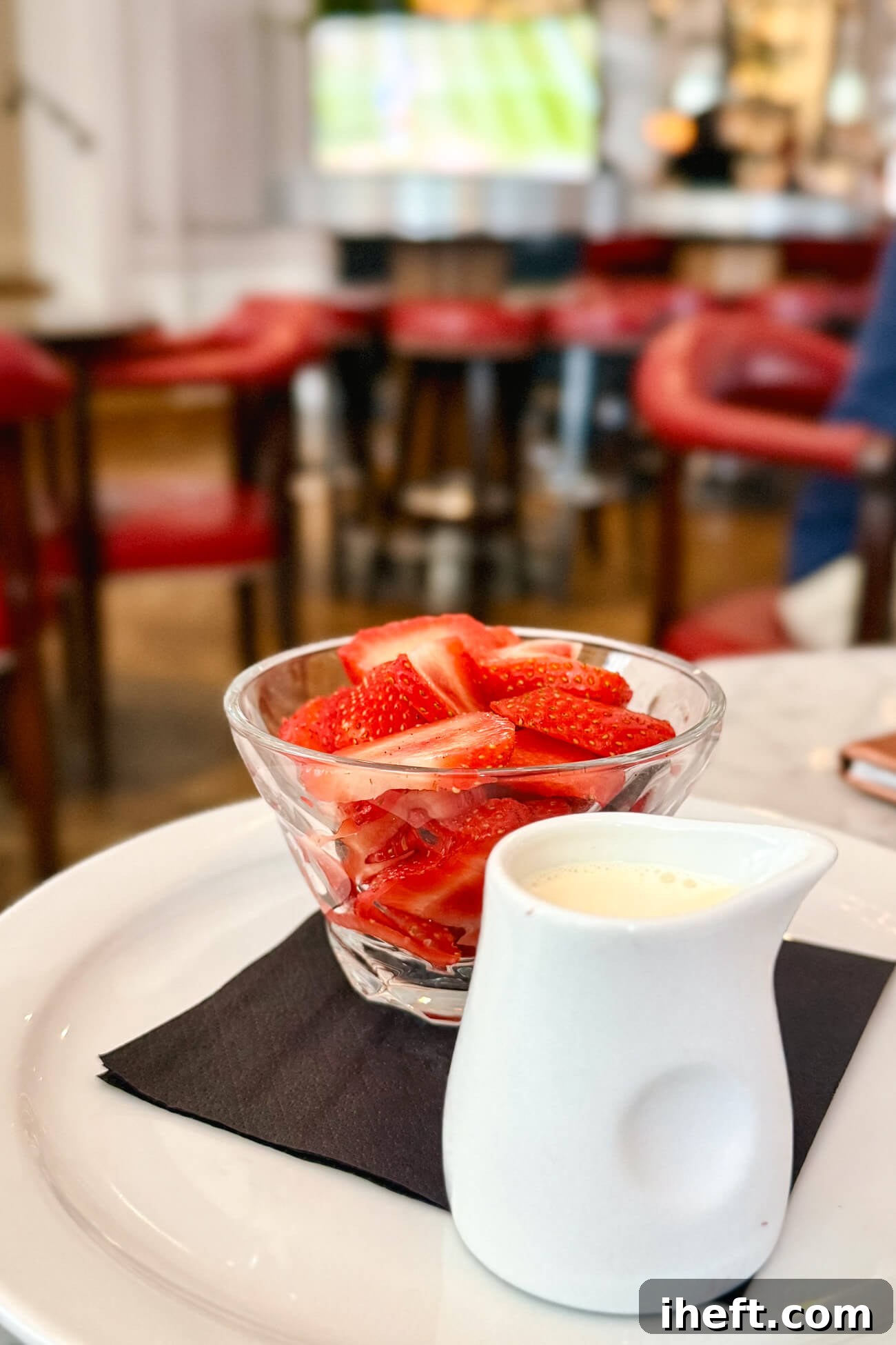 A small glass dish of sliced strawberries with a side of cream four pouring on a restaurant table with tennis on a TV screen in the background. 