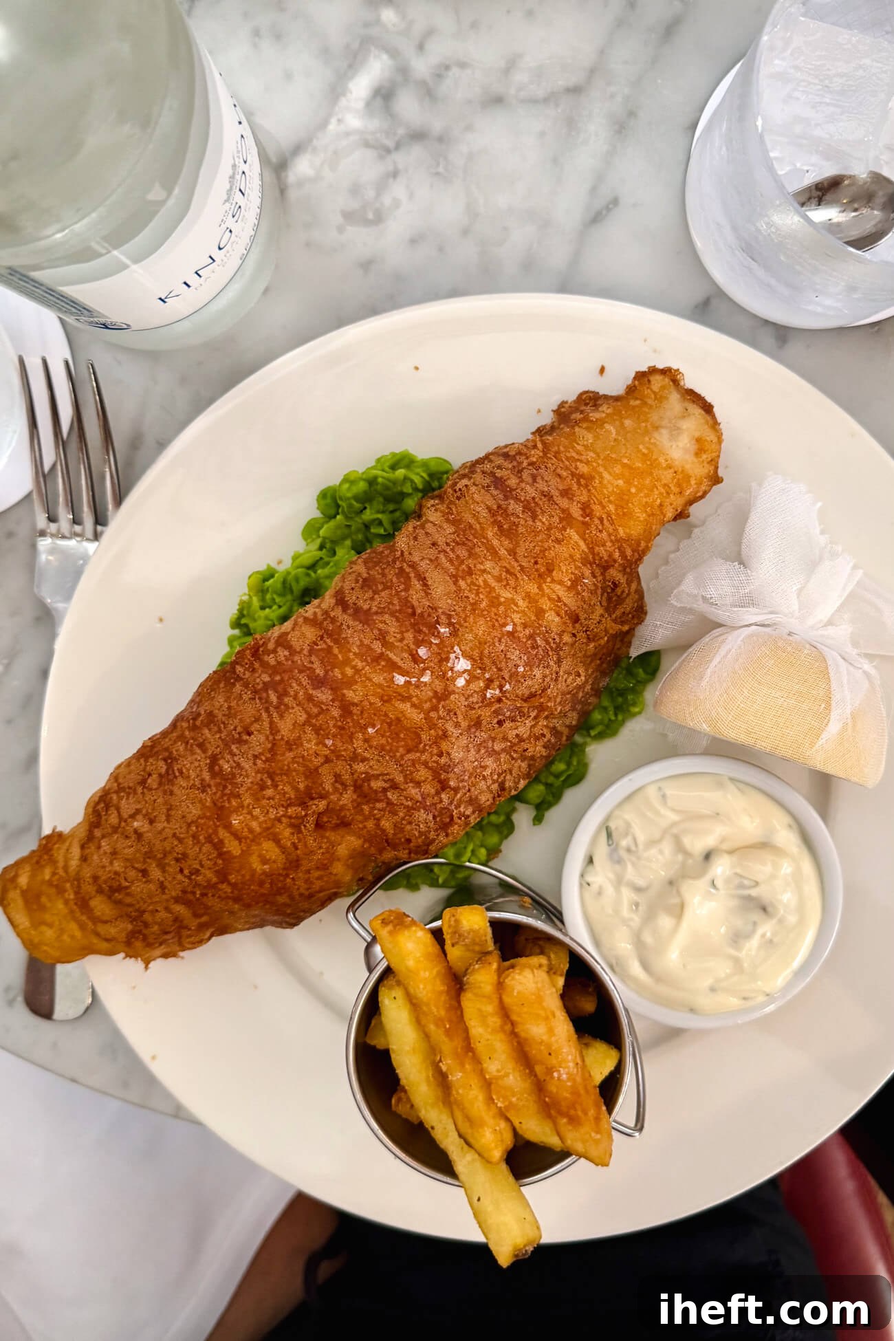An overhead photo of classic British fish and chips made with battered cod over fresh mushy peas served with a small bucket of fries, half a lemon, and tartar sauce.