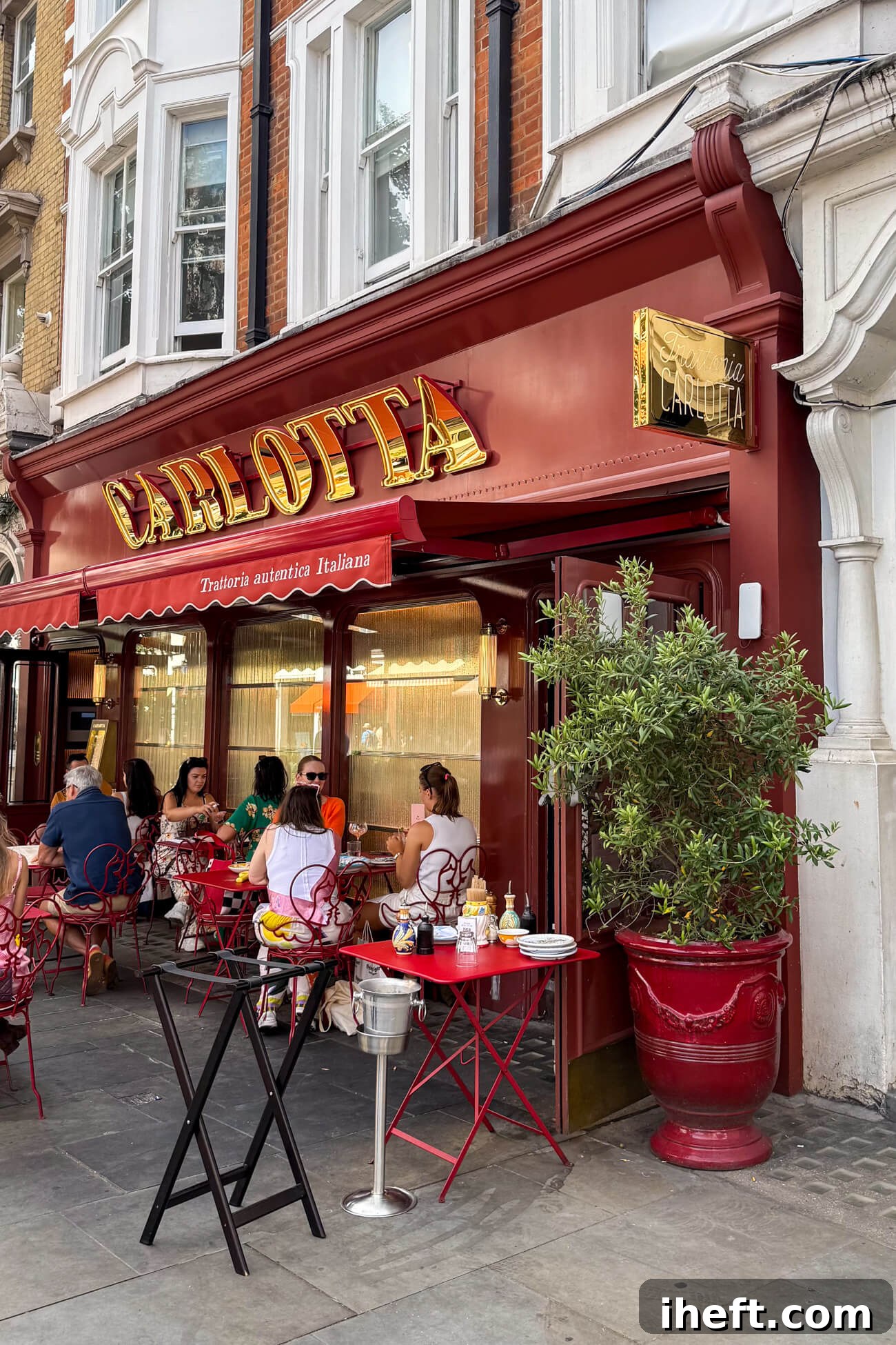 The exterior of Carlotta restaurant in Marylebone, London, featuring a striking red awning with gold lettering. People are enjoying outdoor dining, adding to the vibrant street atmosphere.