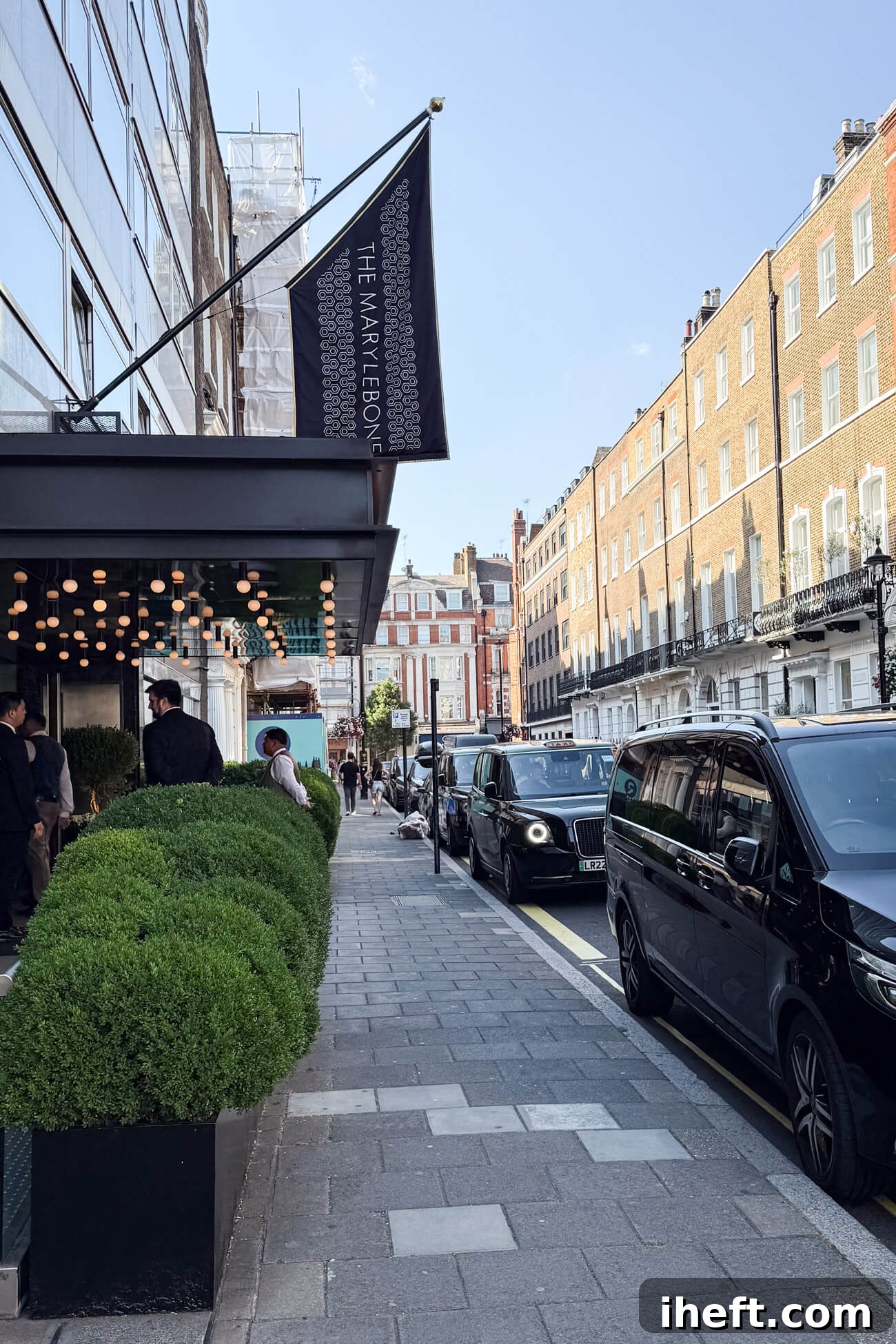 The front of the Marylebone Hotel with black taxis in front. The hotel features a black awning with lights, a black and white flag that says "The Marylebone." Doormen in black suits stand in front. 