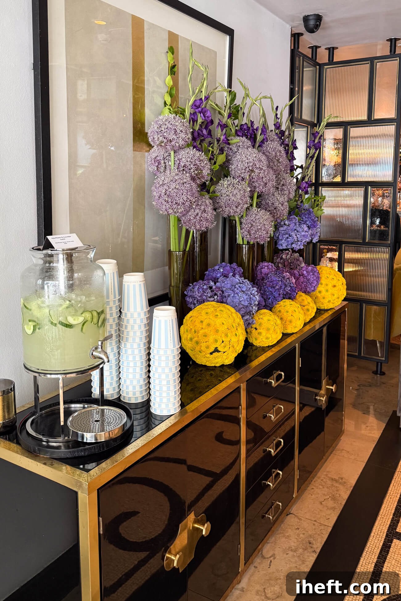 A black and gold buffet table topped with a purple and green flower arrangement and a jug of cucumber beverage with cups in the Marylebone Hotel lobby. 