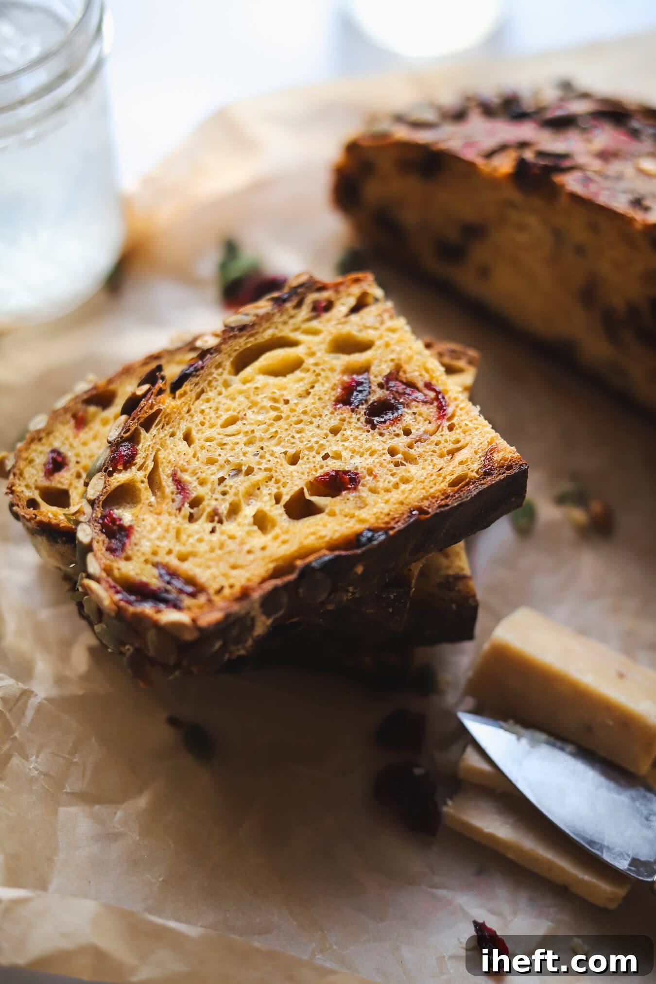 Slices of a seeded pumpkin sourdough bread loaf with dried cranberries and pumpkin seeds on a piece of parchment paper with white cheddar cheese on the side. 