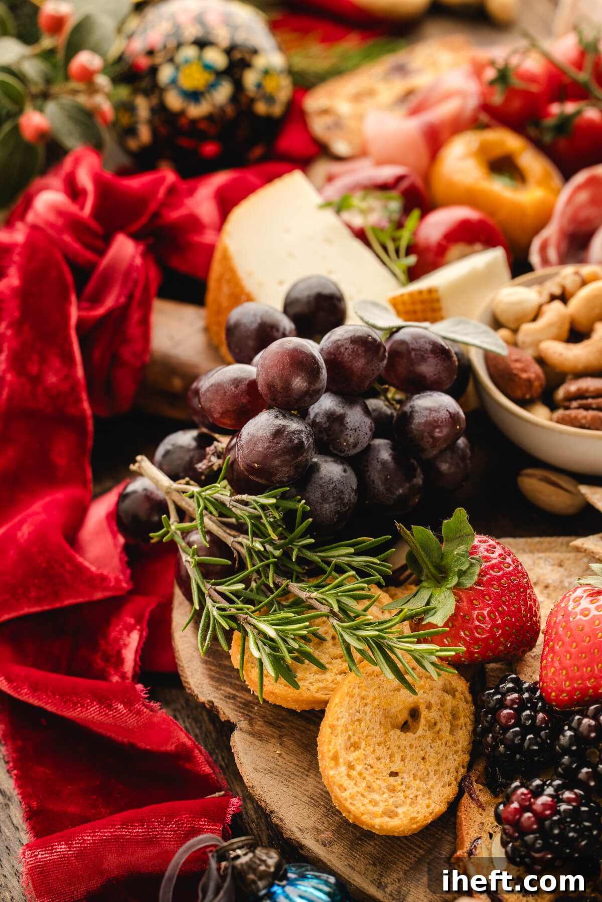 Close-up of Christmas charcuterie board with red velvet ribbon, grapes, strawberries, rosemary, crackers, and festive holiday grazing ingredients.