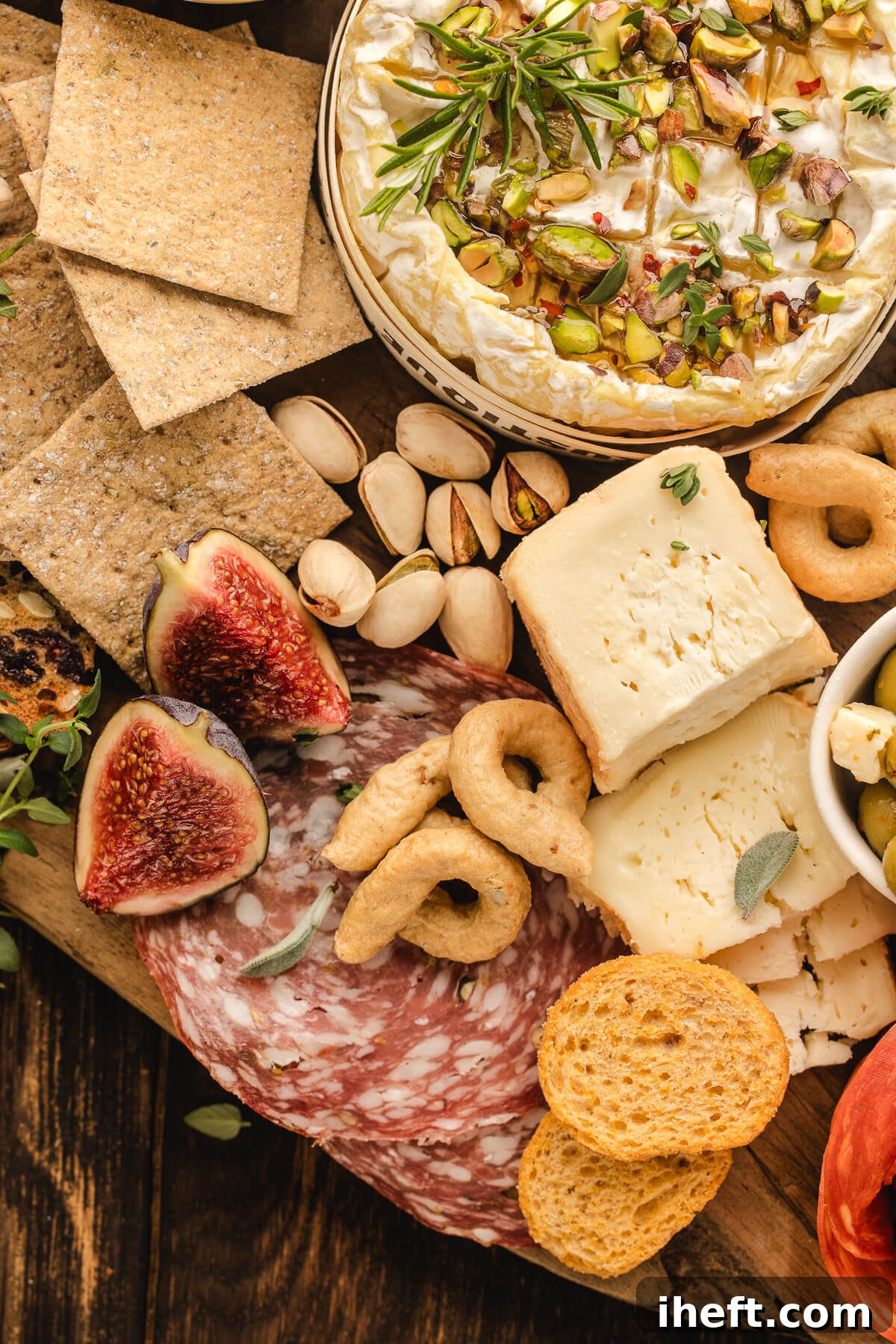 Close-up of a charcuterie board with figs, salami, crackers, pistachios, soft cheese, and whipped brie topped with herbs and nuts.