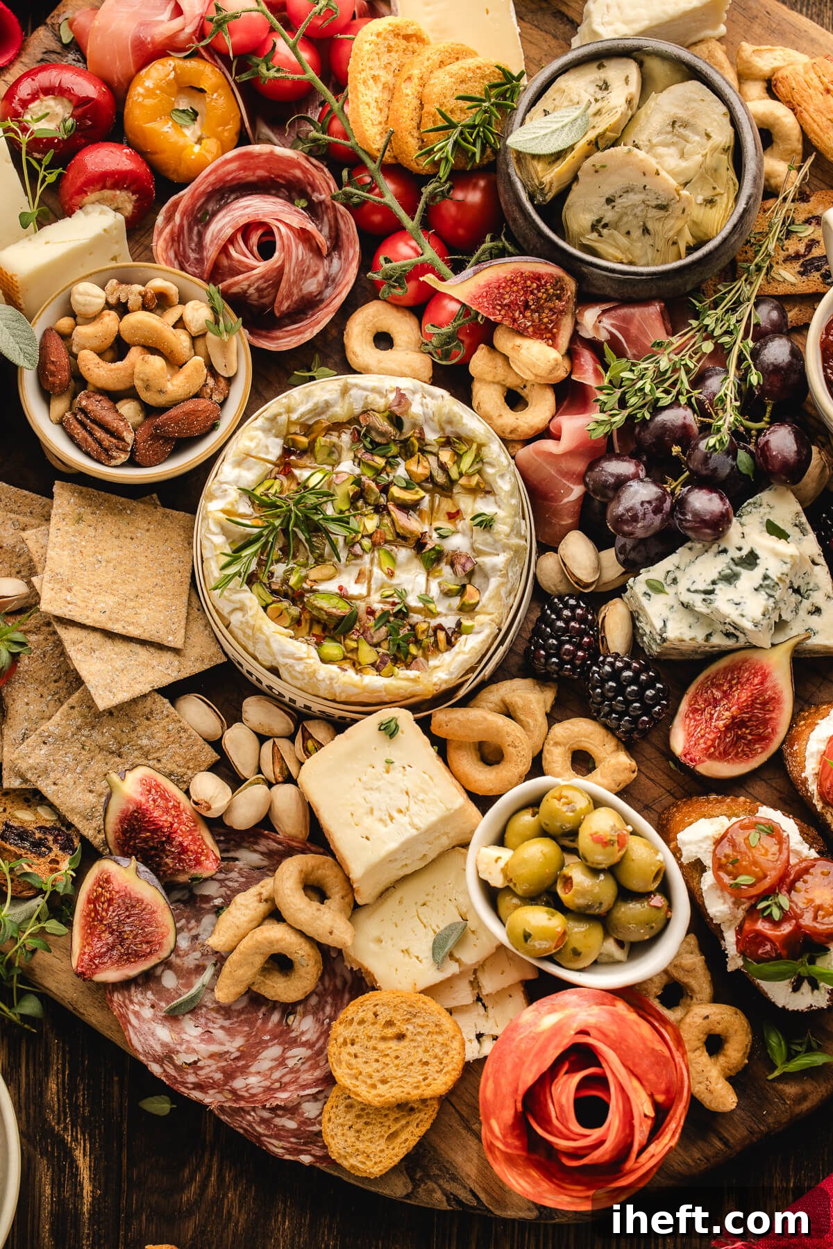 Close-up of a Christmas charcuterie board with baked brie, artichokes, olives, salami, figs, grapes, nuts, crackers, and festive holiday garnishes.