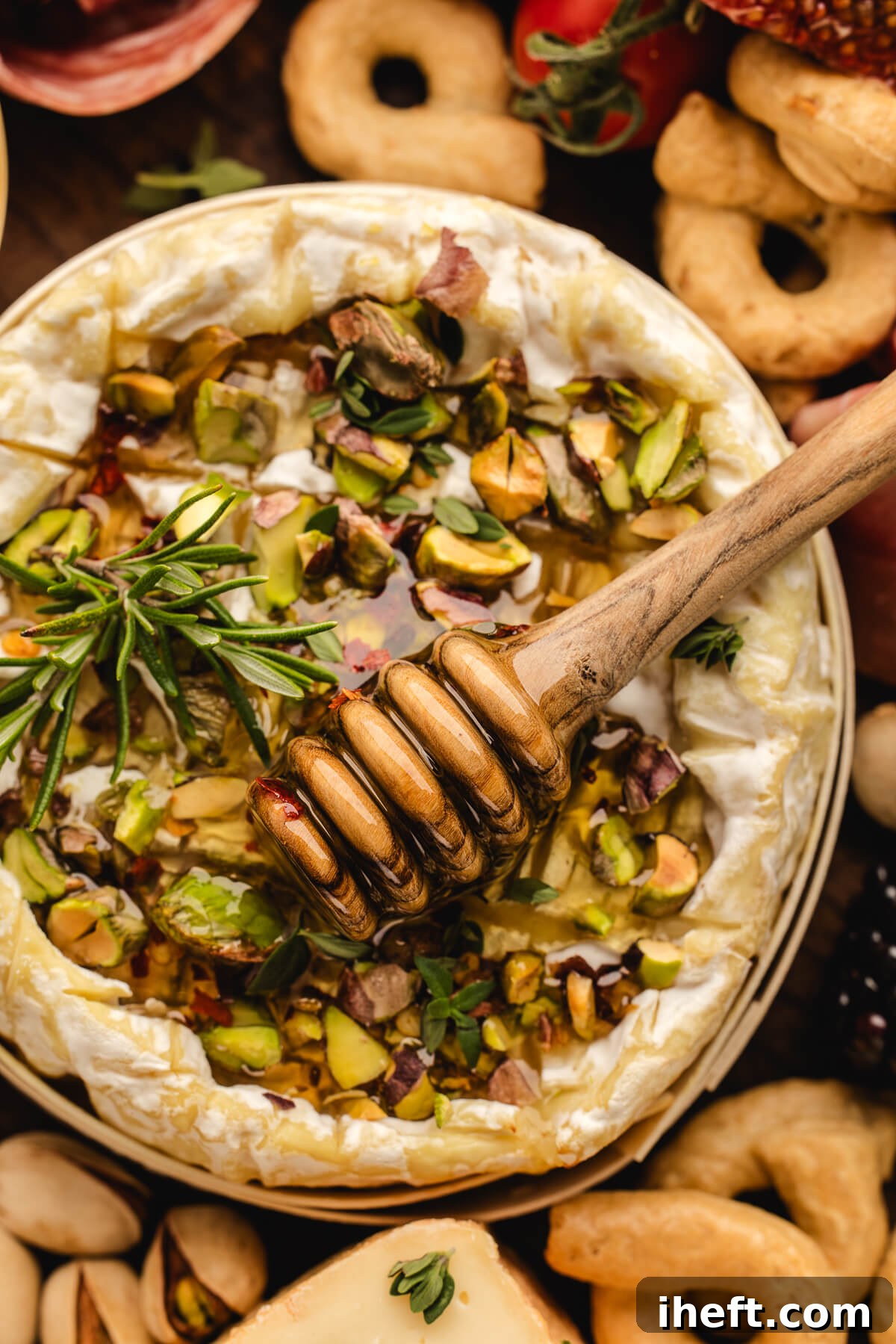 A wheel of baked Brie with honey and pistachios and a wooden honey dipper as part of an appetizer spread. 